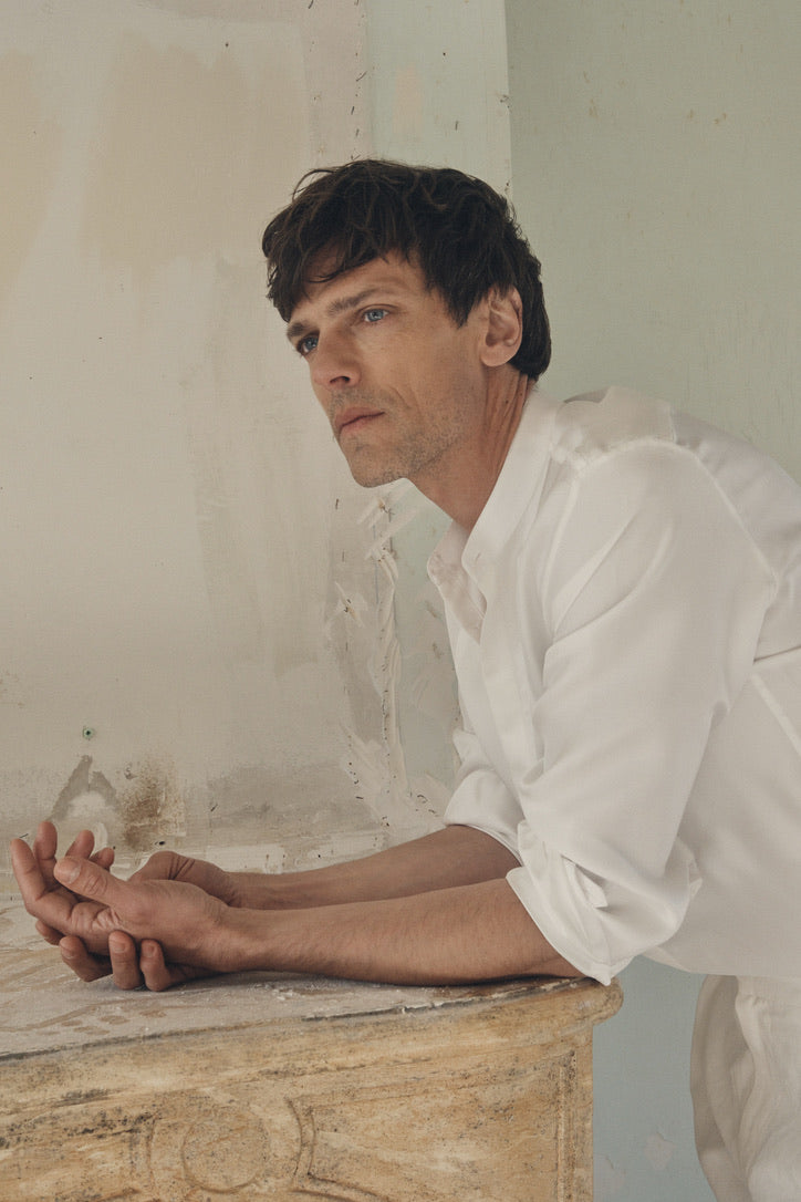 Homme de profil trois-quarts, appuyé en avant sur le rebord d’un vieux manteau de cheminée en pierre patinée, les avant-bras reposant sur le marbre, portant la chemise NOCTURNE en soie blanche à col officier et plastron nid d’abeille. Les manches sont retroussées juste au-dessus des poignets Bourrienne finement froncés, révélant la fluidité satinée du tissu et le mariage subtil de la texture et de la coupe ajustée. En arrière-plan, le mur aux enduits écaillés renforce le contraste entre la délicatesse de la