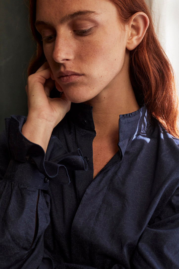 Photo plan poitrine d'une femme qui porte une chemise BOUDOIR bleu marine imprimé liberty de la marque Bourrienne Paris X. La femme rousse à l'air pensive sa main gauche est posée sur sa machoire. On aperçoit le col ancien de la chemise qui se fini d’un liseré de petits volants lui donnant un côté sophistiqué. Les manches bouffantes sont ornées d’un volant ce qui donne un côté original à la chemise. 