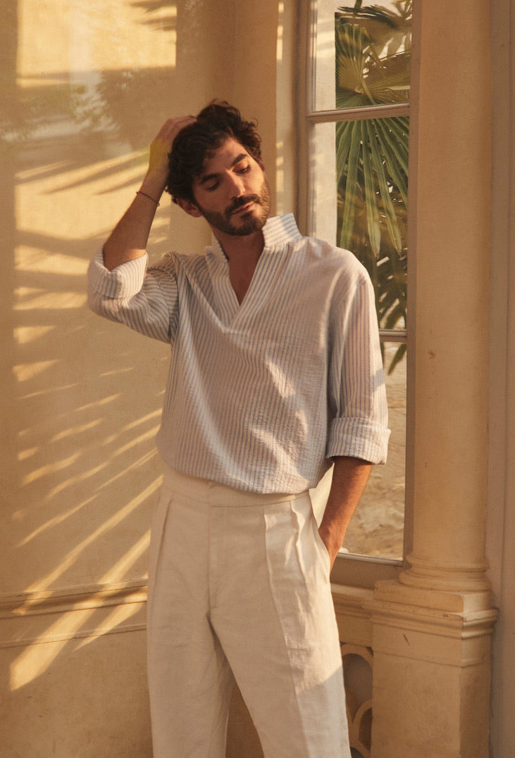 Photo plan américian d'un homme debout,  brun et barbu. Il porte un pantalon en lin blanc et une chemise SCULPTEUR SEERSUCKER dans un léger tissu rayé bleu et blanc. Inspirée des chemises d’artistes et des vareuses, sa coupe ample et sans boutonnage permet de l’enfiler facilement. Son encolure basse lui donne une allure décontractée qui s’adapte à chaque