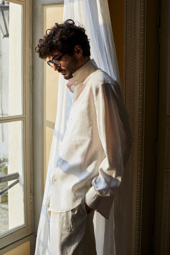 Photo plan américain d'un homme brun barbu de profil. Face à une fenêtre style ancien il porte la chemise OPTIMISTE qui se distingue par son col haut et ses poignets Bourrienne, signatures de la Maison. Confectionnée dans un lin léger de haute qualité, elle offre un confort optimal. Ses deux poches plaquées à l'avant lui apportent une allure structurée. Des broderies discrètes à l'intérieur des poignets et sur le côté ajoutent une touche de détail unique. 