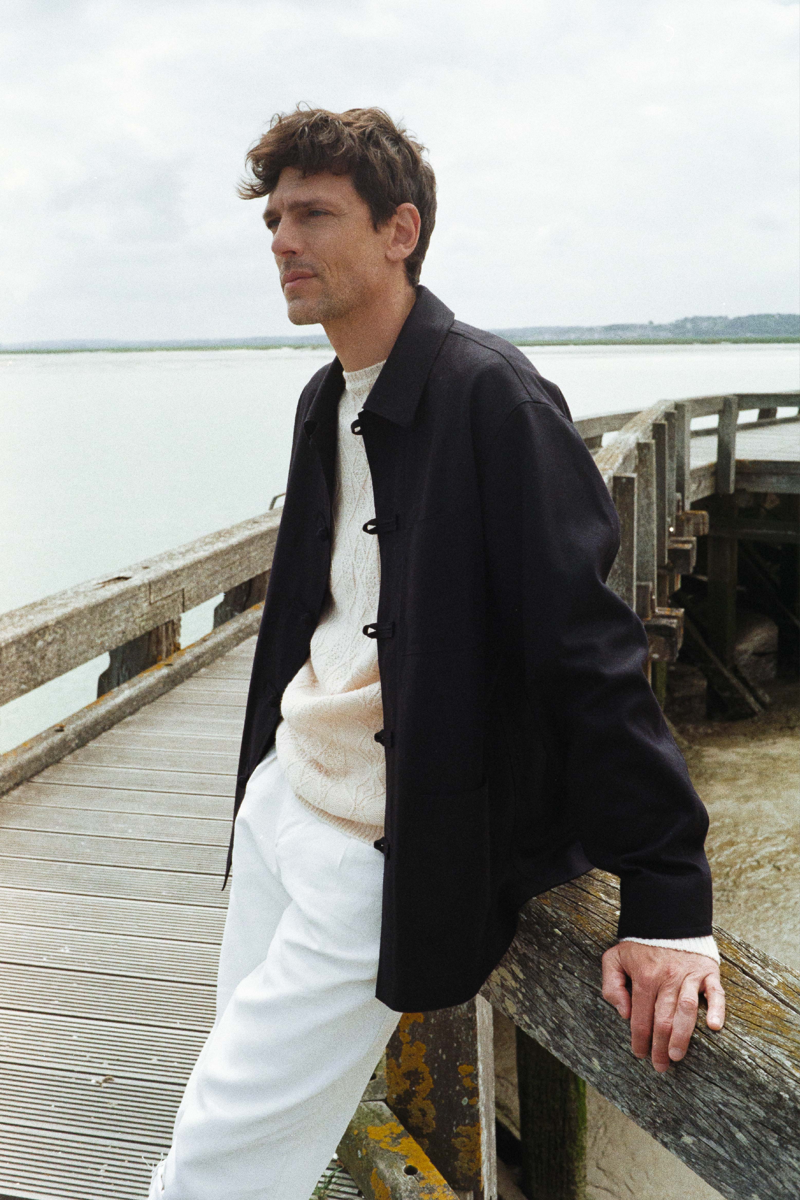 BOURRIENNE PARIS X: Photo prise de profil, le mannequin est sur un ponton au dessus de la mer, il regarde à l’horizon et porte une veste bleu marine à col français, coupe ajustée, poignets classiques, boutonnage travaillé, disposant de trois poches plaquées à l'avant.