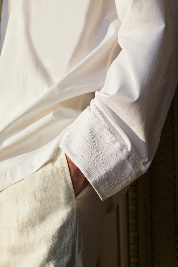 Photo close-up d'une main rentré sur une poche de pantalon, nous voyons une chemise blanche qui se nomme ARTISAN. Des broderies réalisées par un artisan français son présentent sur le col et ses poignets, inspirées par les fresques de l’Hôtel Bourrienne.