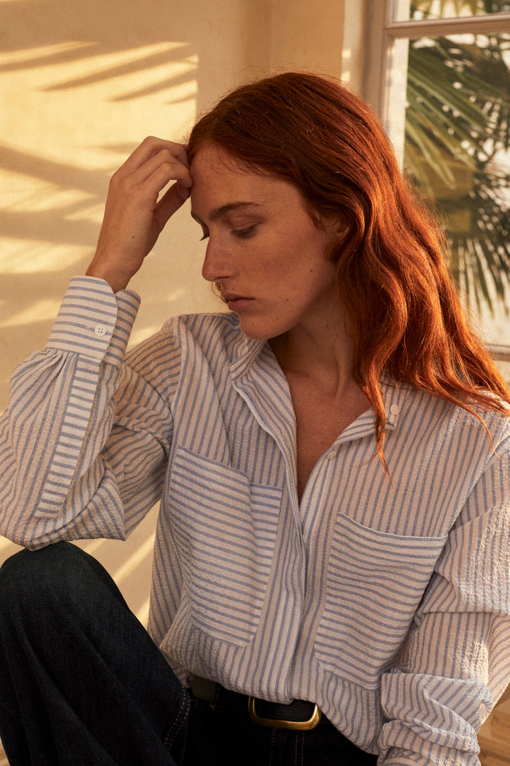 Une photo plan poitrine d'une femme rouse en tailleur. Elle porte une chemise ENCRE SEERSUCKER  un tissu rayé bleu et blanc. Elle a un col haut et des fronces aux manches, deux grandes poches sur le devant. La main droite de la femme est posé sur son front, l'air pensif. 