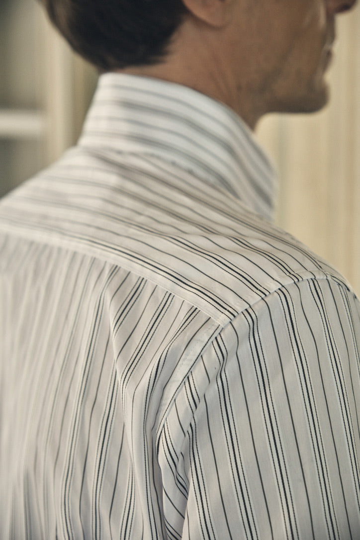 Gros plan sur la nuque et l’épaule droite, où se détache le col montant rigide, boutonné jusqu’au deuxième cran, ainsi que la découpe nette de l’empiècement dos.
Les fines rayures kaki et gris, régulières et parallèles, courent sur une popeline blanc cassé d’aspect légèrement satiné.
Un discret pli creux sous l’empiècement souligne la coupe ajustée et offre une souplesse subtile au dos de la chemise.
La lumière tamisée met en valeur la finesse des surpiqûres et la qualité du tombé du tissu.