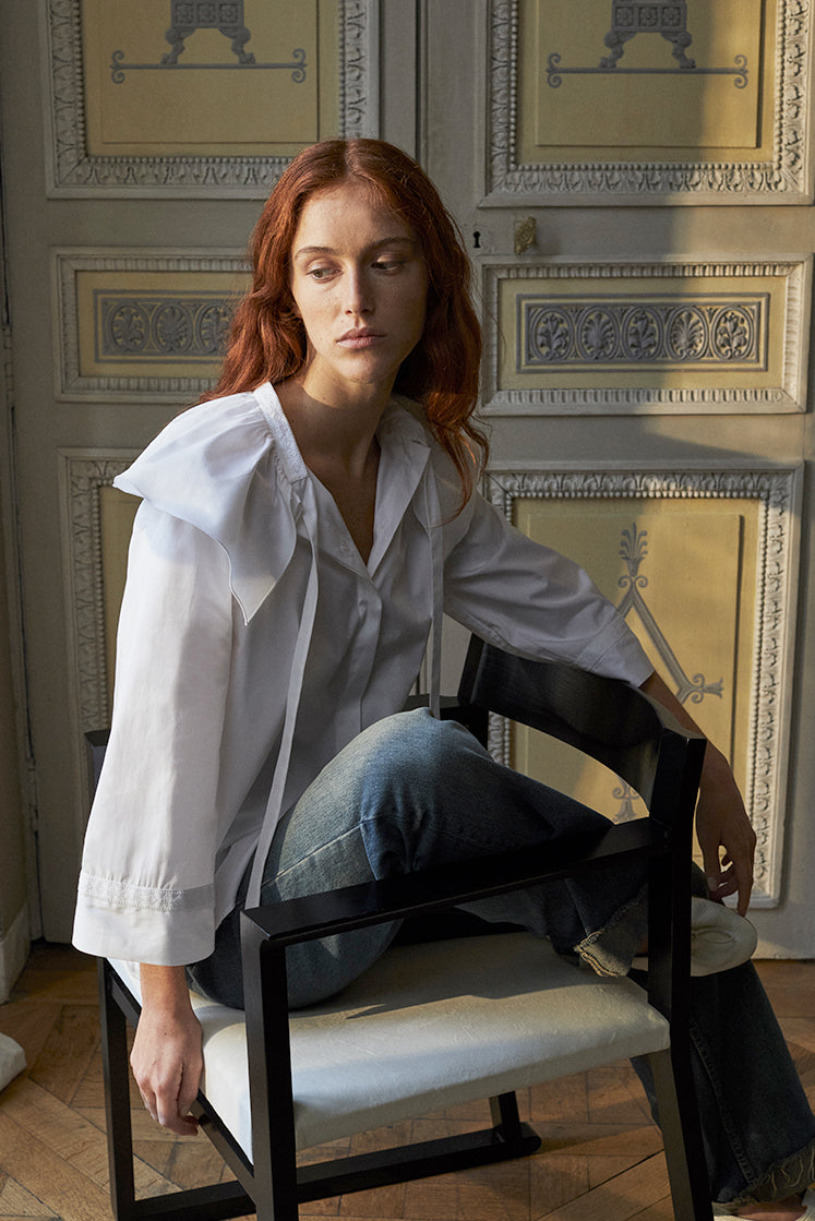 Mannequin rousse, assise sur une chaise, portant la chemise Alizé de Bourrienne Paris X avec un col brodé et orné d'un volant assumé, des maches évasées coupées trois-quart également brodées sur les poignets.