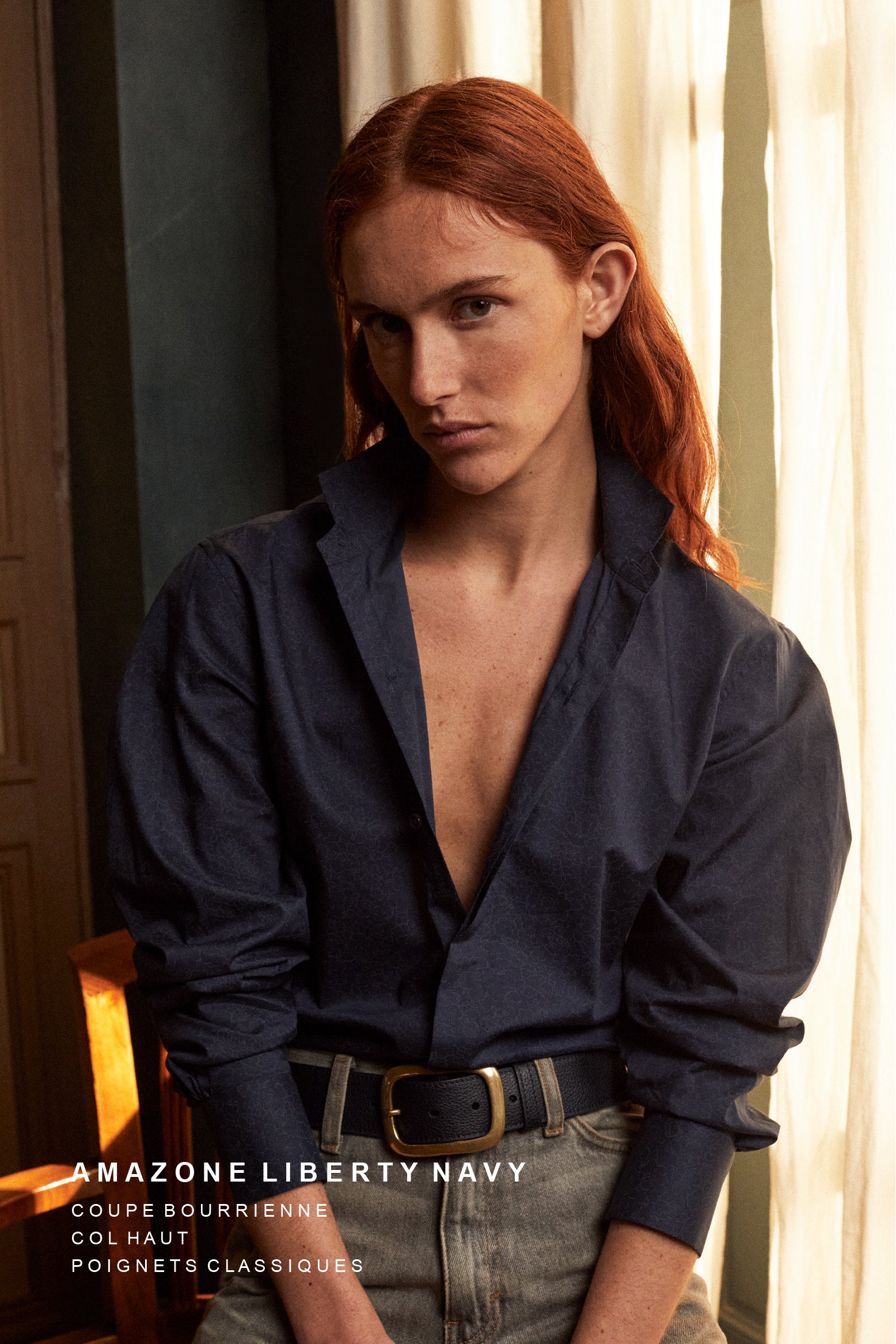 Photo plan américain, une femme rousse est légèrement assise sur une chaise, elle regarde l'objectif. Elle porte une chemise AMAZONE bleu marine imprimé Liberty Bourrienne Paris X. Ses manches sont légèrement remontées, les manches des épaules sont légèrement bouffantes avec un col ancien qui revisite le vestiaire homme. 