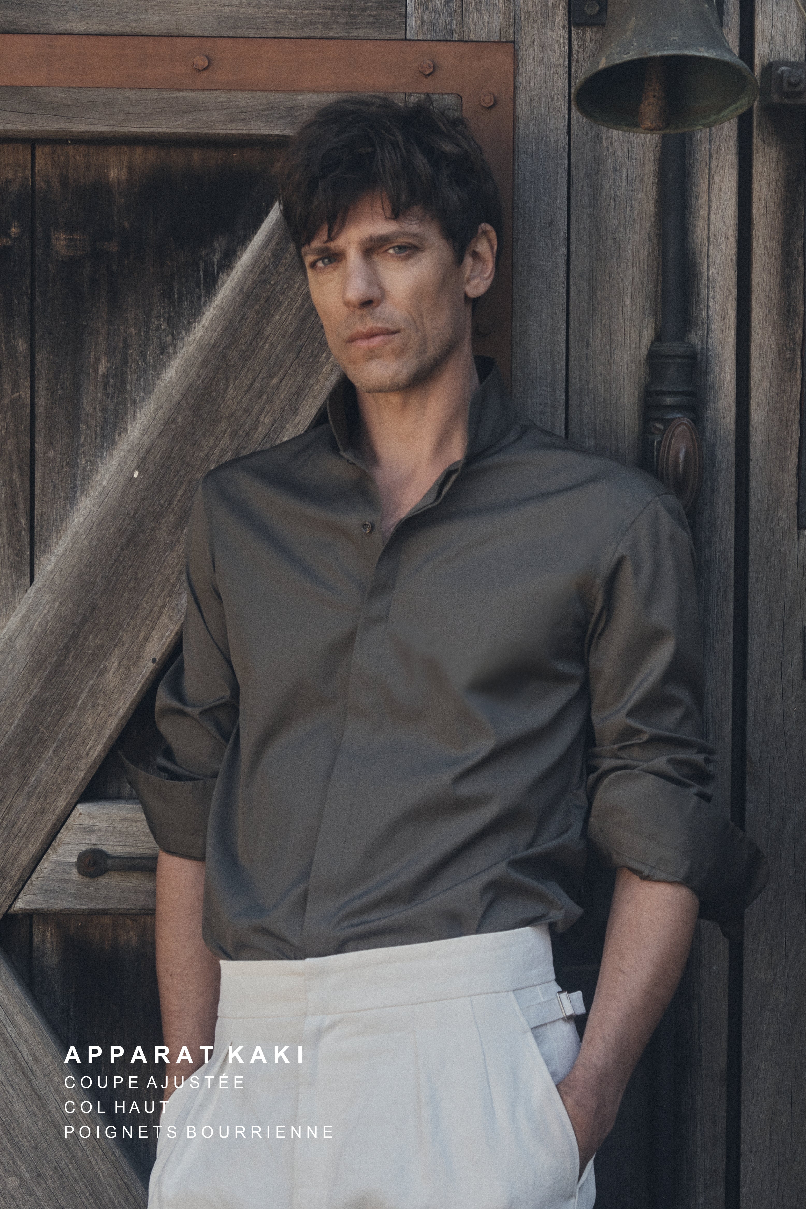 Un homme de trois-quarts pose contre une porte en bois patiné, main droite dans la poche de son pantalon blanc cassé. Il porte une chemise ajustée kaki foncé en popeline, col montant fermé au deuxième cran et manches retroussées. Le contraste entre le haut sombre et le bas lumineux souligne la modernité de la coupe et la qualité du tissu.