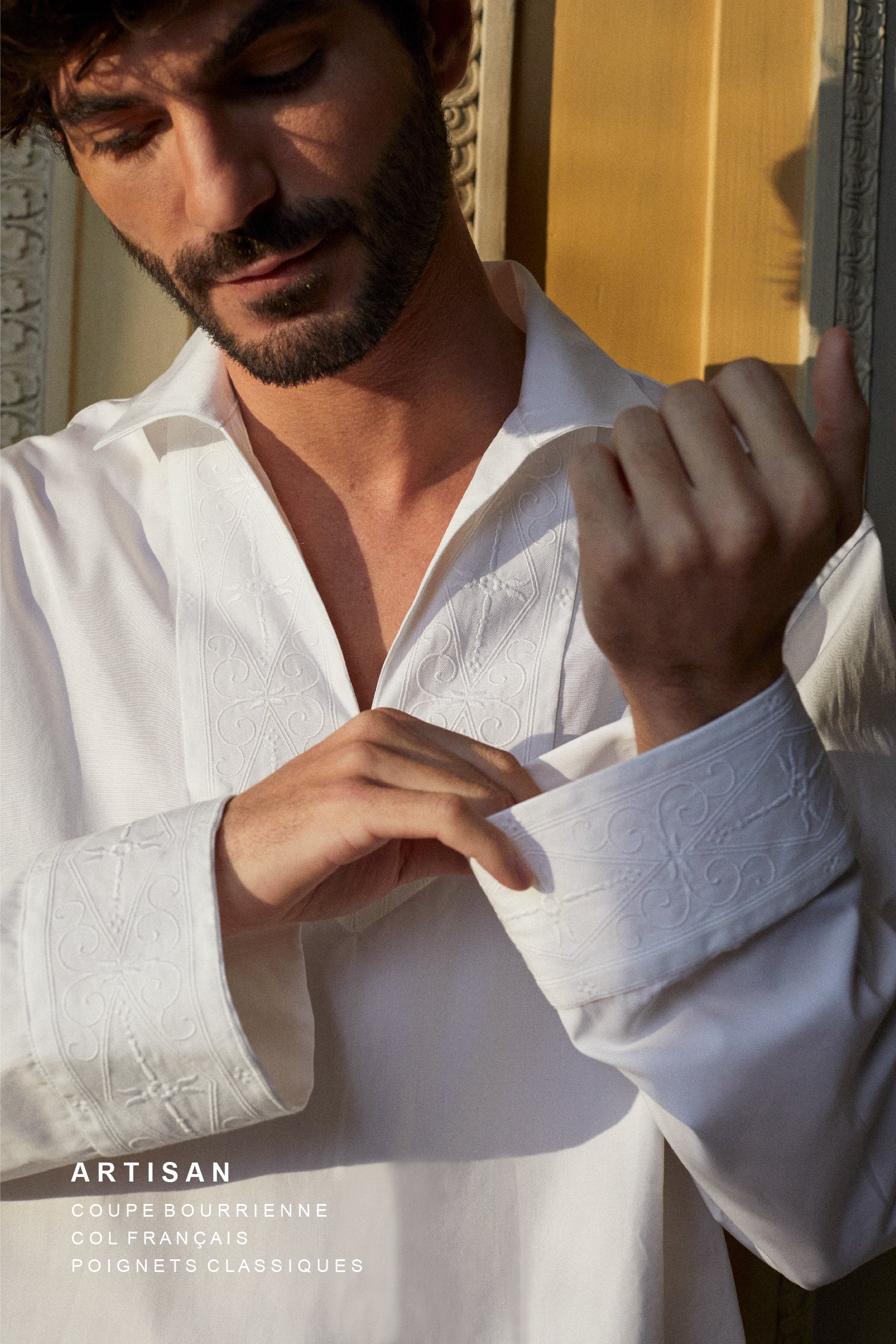 Photo plan poitrine d'une homme brun et barbu, il porte une chemise blanche avec un col V, la chemise se nomme ARTISAN. L'homme croise ses mains, des broderies réalisées par un artisan français son présentent sur le col et ses poignets, inspirées par les fresques de l’Hôtel Bourrienne.