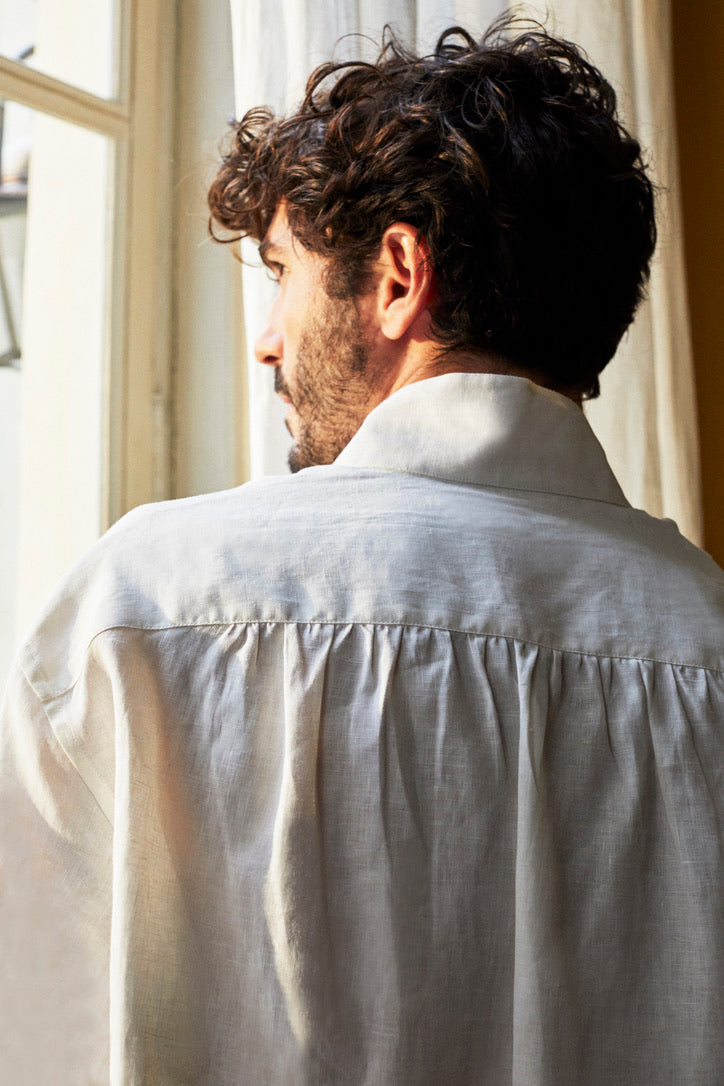 Photo gros plan d'un homme brun, barbue, de dos, portant une chemise nommée OPTIMISTE. Elle se distingue par son col haut et ses poignets Bourrienne, signatures de la Maison. Confectionnée dans un lin léger de haute qualité, elle offre un confort optimal. Ses deux poches plaquées à l'avant lui apportent une allure structurée. Des broderies discrètes à l'intérieur des poignets et sur le côté ajoutent une touche de détails uniques. 