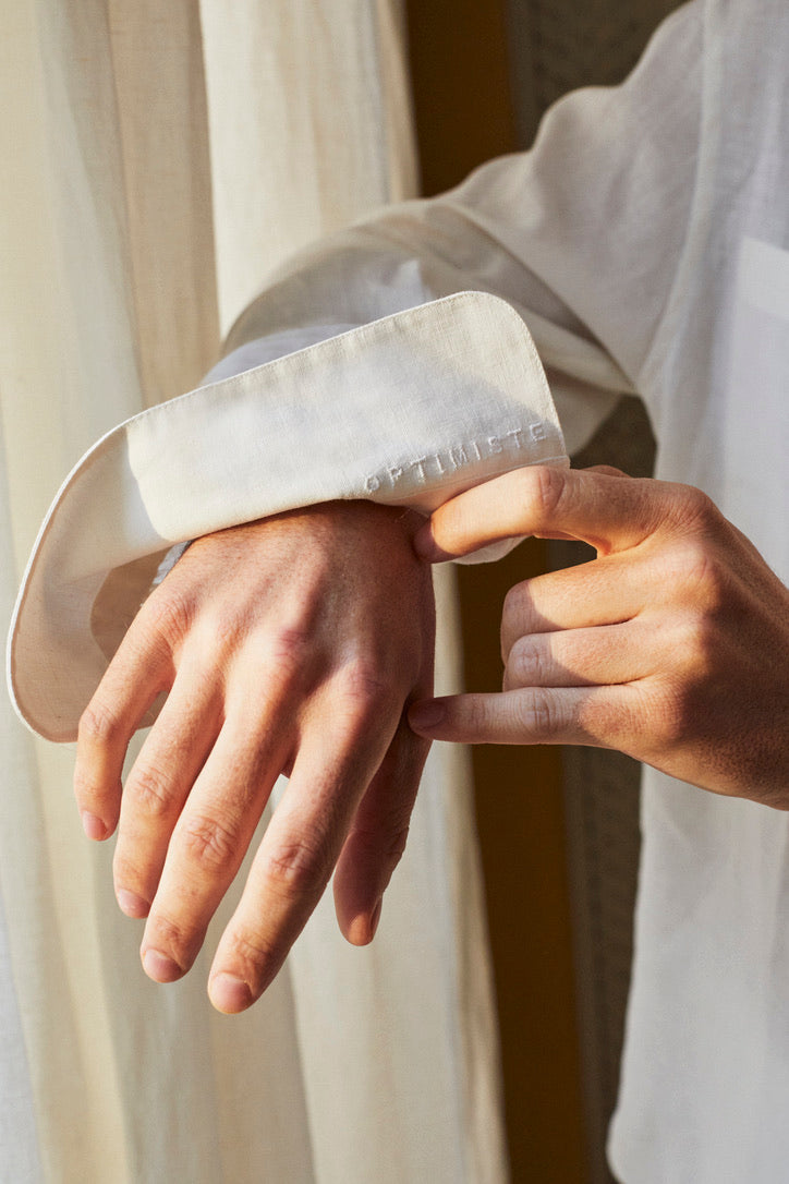 Photo gros plan de la chemise OPTIMISTE . On y voit les mains du mannequin, qui retournent le poignet de sa chemise laissant apparaitre une broderie: OPTIMISTE: le nom de la chemise. Des broderies discrètes à l'intérieur des poignets et sur le côté ajoutent une touche de détails uniques. 