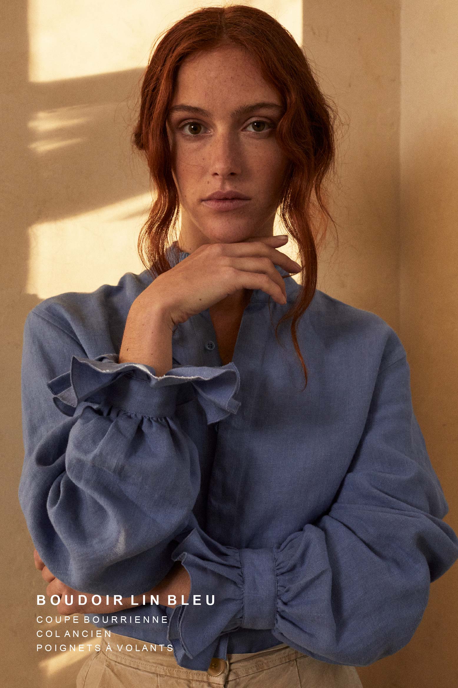 Femme rousse portant une chemise en lin bleu BOUDOIR de Bourrienne Paris X, avec col ancien et poignets à volants. Elle pose dans une lumière dorée, projetant des ombres élégantes sur le mur.