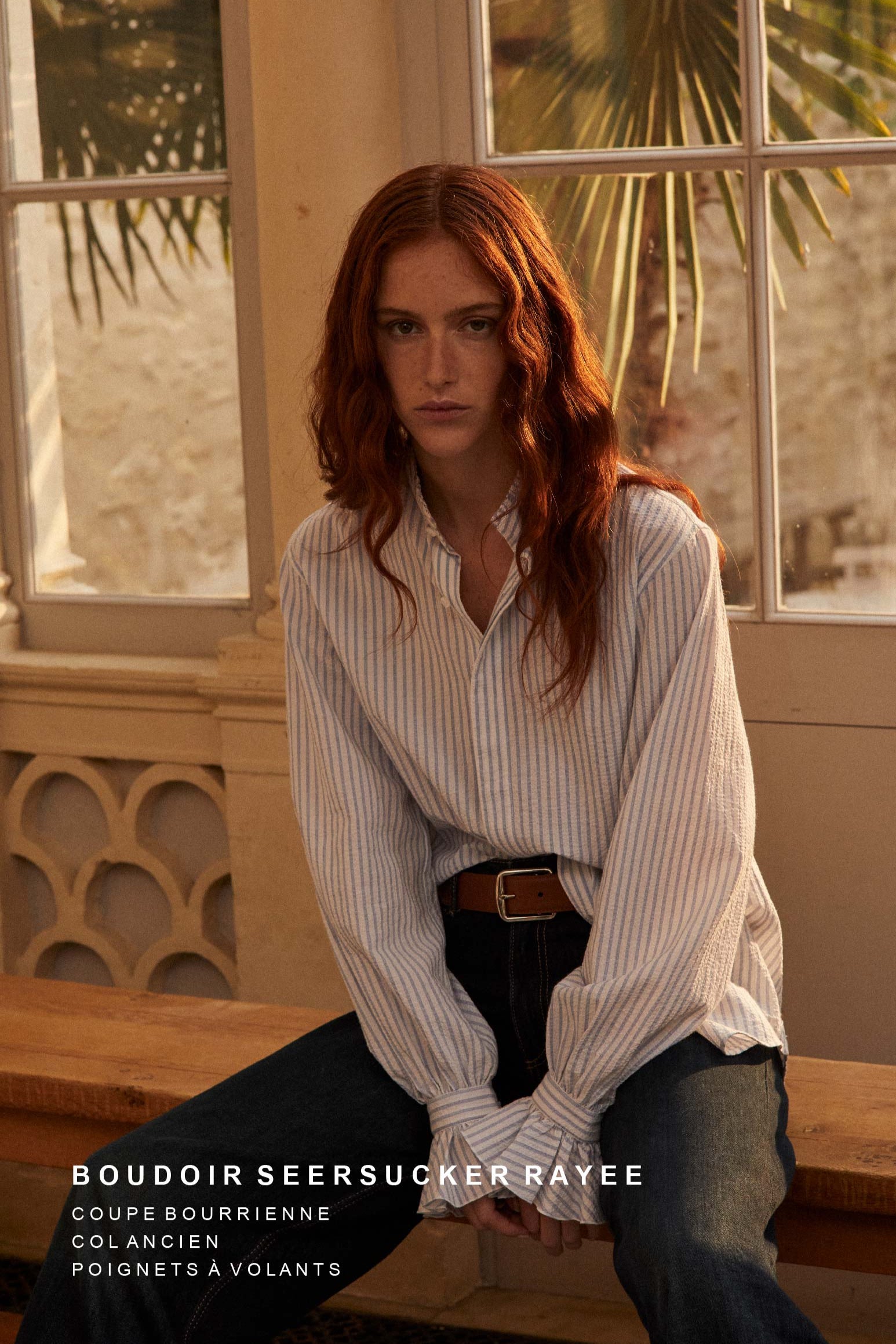 Photo plan américain d'une femme rousse avec les cheveux détachés, elle est asssise sur un banc en bois. Elle porte une chemise Boudoir Seersucker Rayée Bleu qui est rentré sous un jean taille haute, elle est soulignée par une ceinture en cuir marron. 