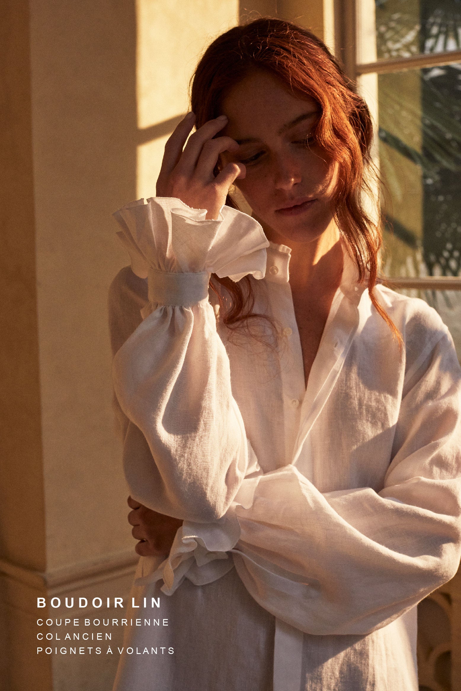 Photo plan américain d'une femme rousse portant un chignon, deux mèches tombantes sur son visage. Elle porte une chemise BOUDOIR dans un lin blanc plein de détails raffinés : un col haut subtilement bordé de petits volants pour une touche de sophistication et des manches légèrement bouffantes ornées de volants, qui tombent avec élégance sur la main. Sa main touche délicatement son visage, l'air pensive. La chemise confectionnée dans un lin belge de Libeco. 