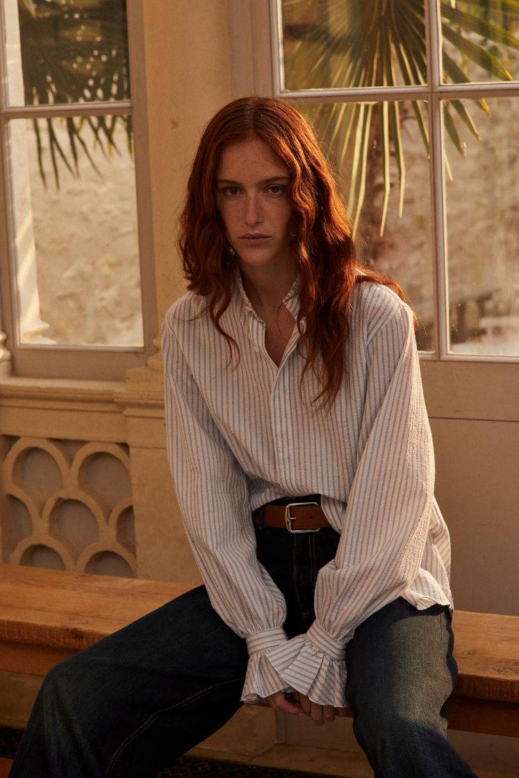 Photo plan américain d'une femme rousse avec les cheveux détachés, elle est asssise sur un banc en bois. Elle porte une chemise Boudoir Seersucker Rayée Bleu qui est rentré sous un jean taille haute, elle est soulignée par une ceinture en cuir marron. 