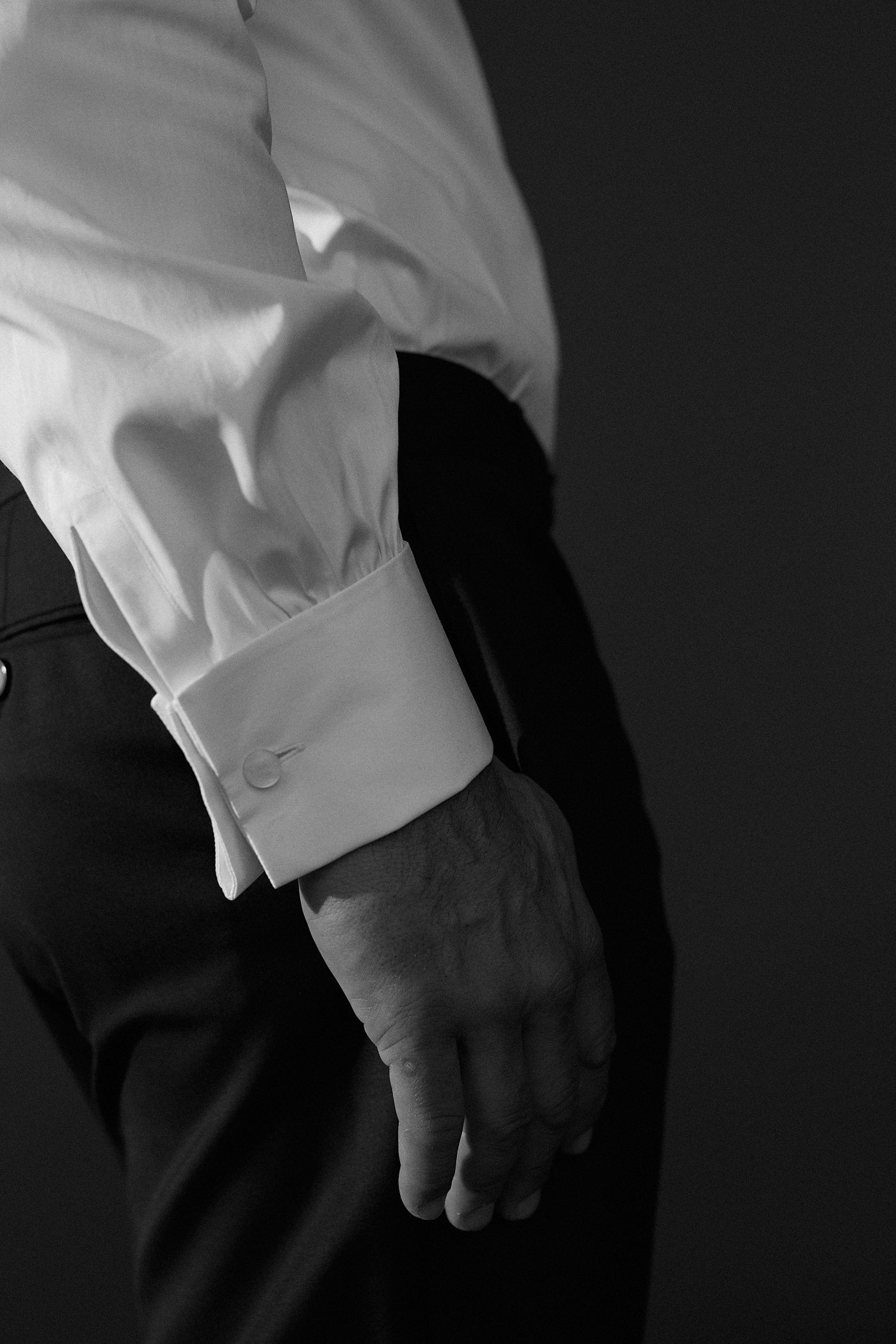 Photo noir et blanc plan serré d'un poignet d'homme, il porte une chemise en popeline blanche avec un poignet mousquetaire, on voit les fronces sur le debut du poignet et la bouton qui ferme le poignet. 