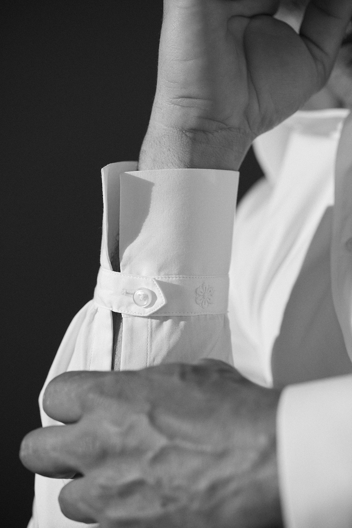 Photo en noir et blanc gros plan, d'une chemise blanche en popeline double retors. Au premier plan la main gauche d'un homme qui touche son avant bras droit, on aperçoit très visiblement  le poignet Bourrienne avec la fleur Bourrienne brodée. 