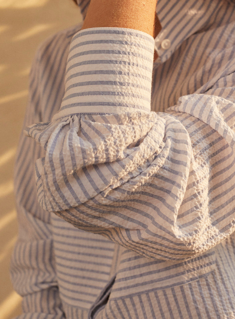 Une photo gros plan d'un avant-bras. Nous voyons de près la chemise ENCRE SEERSUCKER dans un tissu rayé bleu et blanc. Elle a un col haut et des fronces aux manches, deux grandes poches sur le devant.