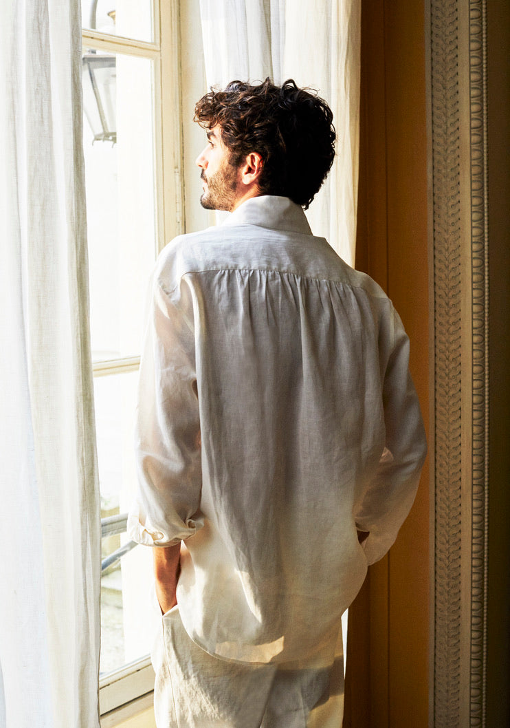 Photo plan américain d'un homme de dos, les mains dans les poches, il porte la chemise OPTIMISTE. Une chemise qui se  distingue par son col haut et ses poignets Bourrienne, signatures de la Maison. Confectionnée dans un lin léger de haute qualité, elle offre un confort optimal. Ses deux poches plaquées à l'avant lui apportent une allure structurée. Des broderies discrètes à l'intérieur des poignets et sur le côté ajoutent une touche de détails uniques. 