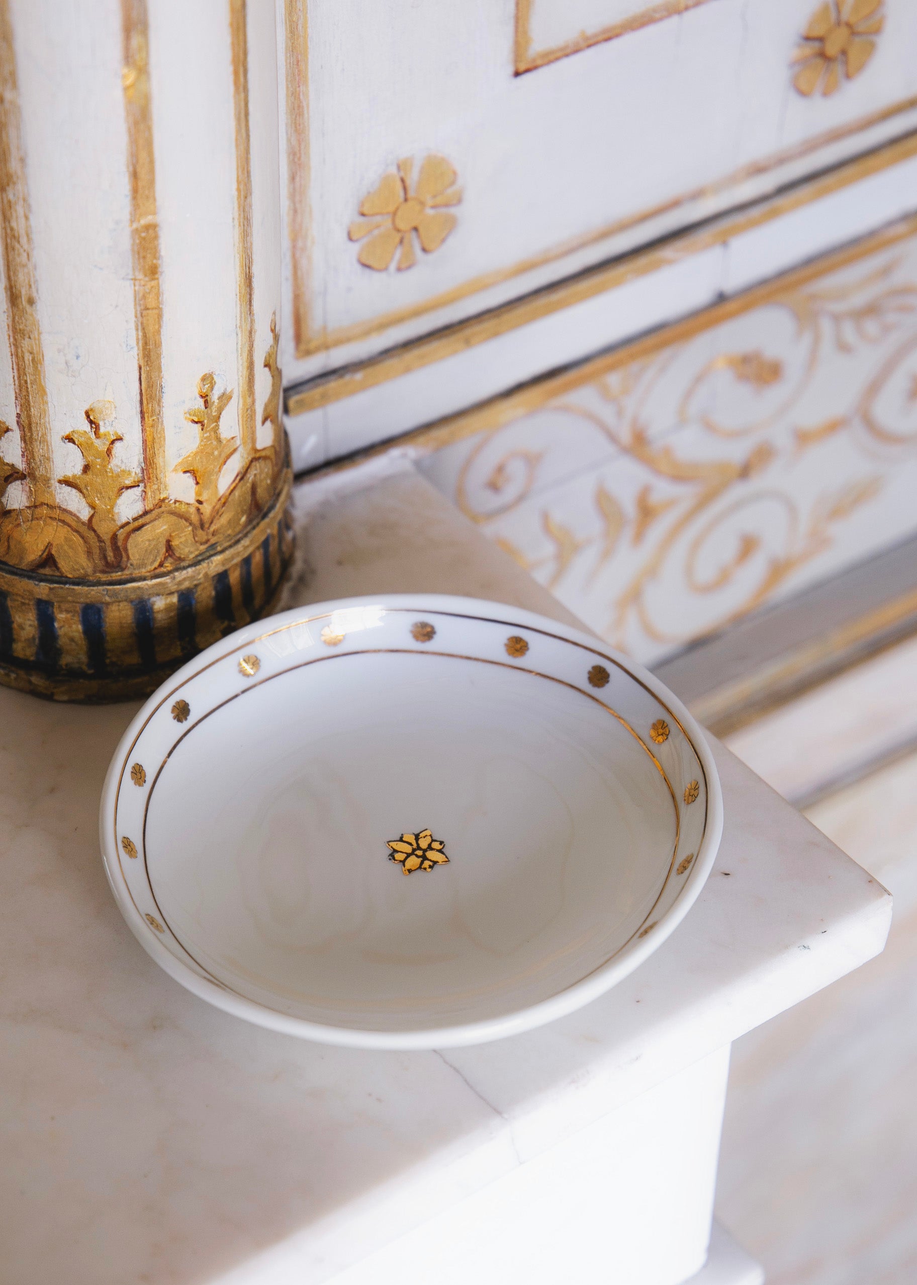 Coupelle en porcelaine blanche ornée de motifs dorés posée sur un coin de cheminée dans l'Hôtel de Bourrienne. On peut apercevoir sur les murs le motif de la collection inspiré des fresques de l'Hôtel.