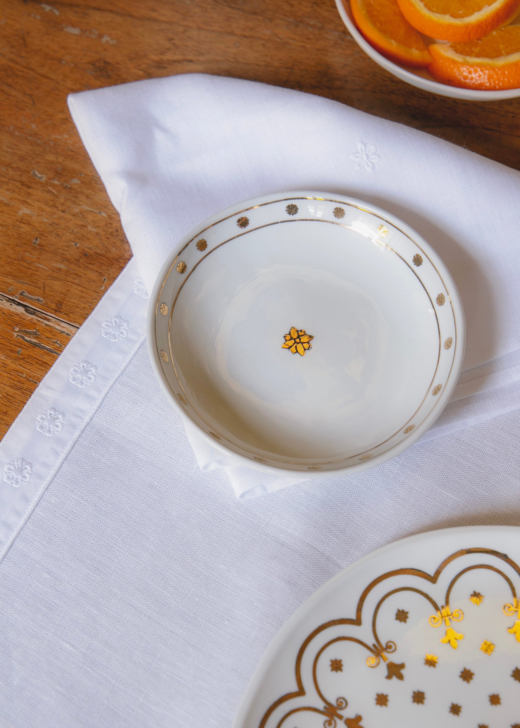 Coupelle en porcelaine blanche ornée de motifs dorés reprenant la fleur de l'Hôtel de Bourrienne, posée sur une serviette blanche et un set de table blanc ornés du même motif.