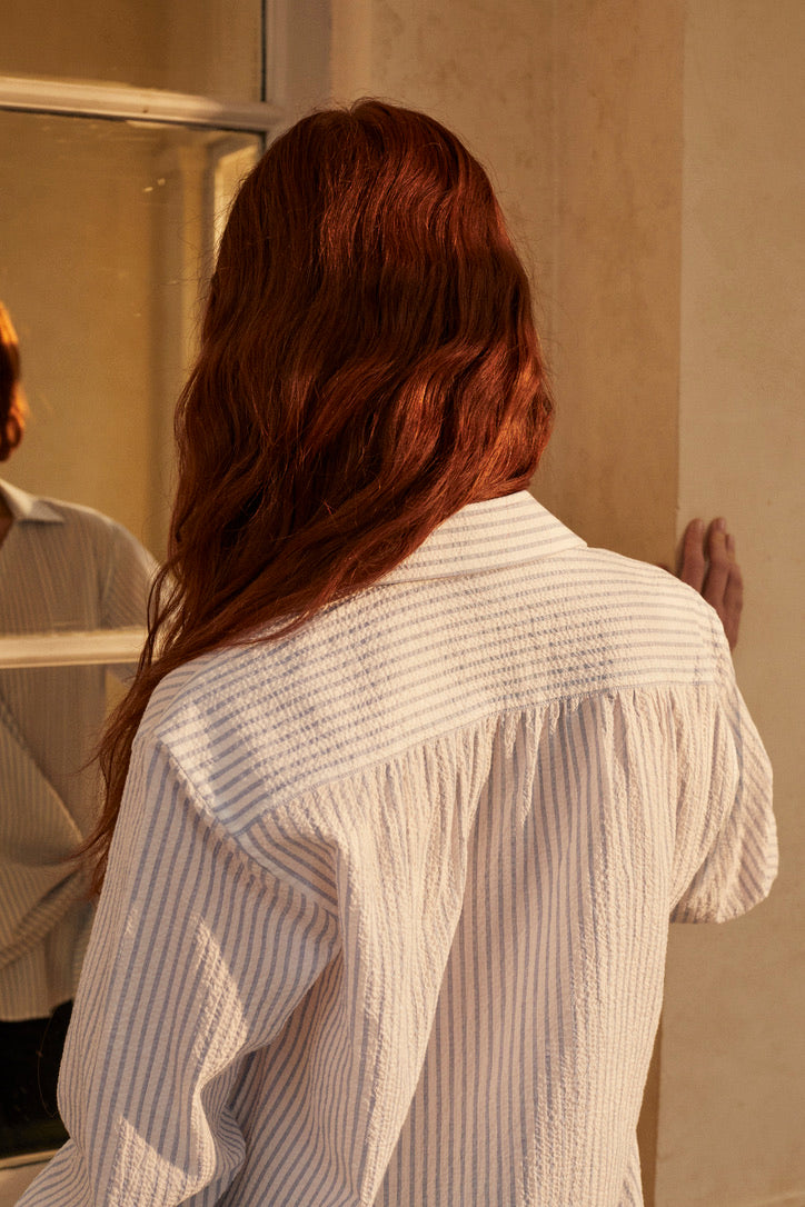 Photo plan taille, d'une femme debout de dos, les cheveux détachés tombant vers la gauche. Sa main droite est posée sur un mur. Elle porte une chemise SAUVAGE SEERSUCKER s'inspire des coupes cache-cœur et se distingue par son tombé fluide et élégant. Sa coupe ample et sa matière légère à fines rayures bleues en font une pièce idéale pour l'été. Son pan supérieur, glissé dans un passant, crée un effet cache-cœur 