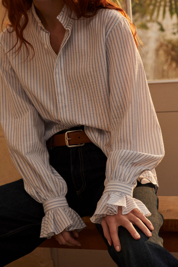 Photo plan poitrine  d'une femme rousse  assise sur un banc en bois. Son visage est coupé, elle porte une chemise Boudoir Seersucker Rayée Bleu qui est rentrée sous un jean taille haute, elle est soulignée par une ceinture en cuir marron. Ses mains sont posées sur son jean, nous apercevons plus en détails les volants des poignets, signature de la Maison Bourrienne. 