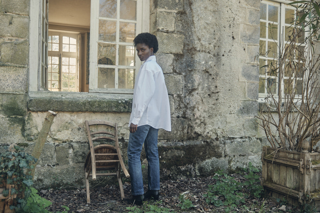 Femme portant la chemise MERVEILLEUSE, posant devant la fenêtre d'une ancienne maison.