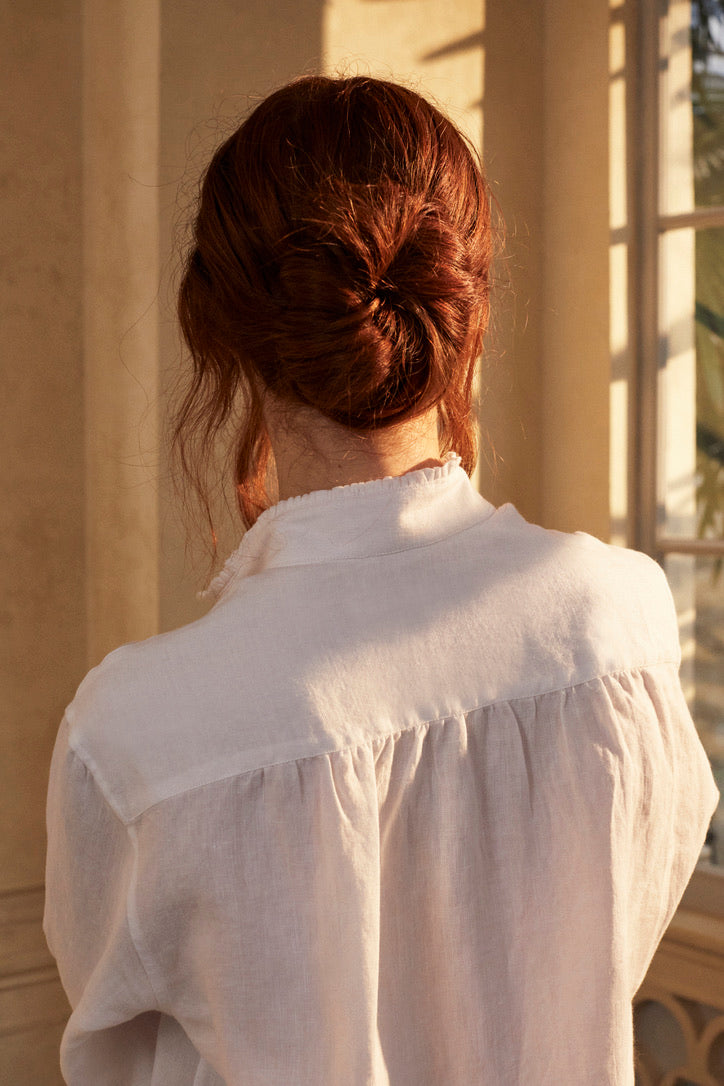 Photo plan poitrine d'une femme rousse de dos avec un chignon. Elle porte une chemise BOUDOIR en LIN Blanc aérien et léger. Des détails raffinés sur le col haut subtilement bordé de petits volants et des manches légèrement bouffantes ornées de volants, qui tombent avec élégance sur la main. 