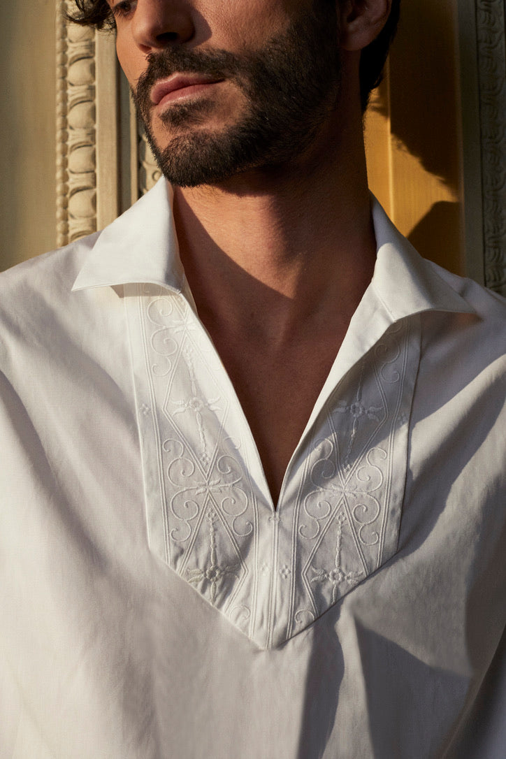 Photo close up d'une homme brun barbu, il porte une chemise blanche avec un col V, la chemise se nomme ARTISAN. La coupe Bourrienne est ample et épurée, des broderies réalisées par un artisan français son présentent sur le col et ses poignets, inspirées par les fresques de l’Hôtel Bourrienne.