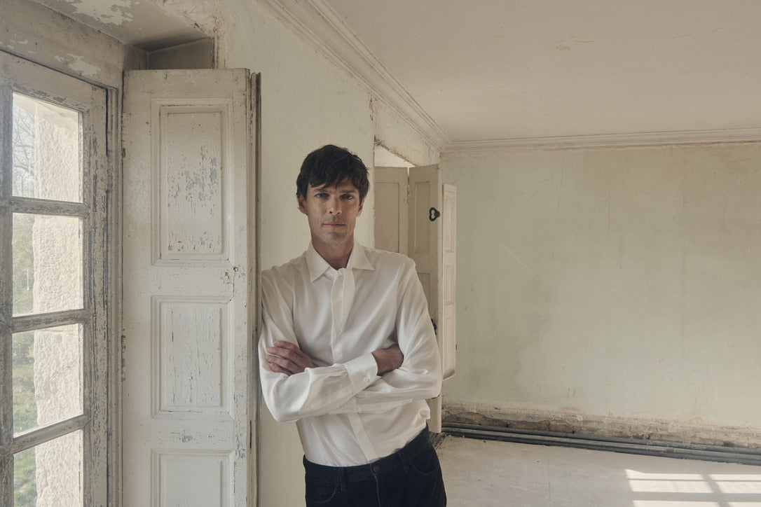 Homme de face, bras croisés et appuyé contre un volet ancien, vêtu de la chemise PALETTE en soie blanche à coupe Bourrienne et col français noué en large nœud décontracté. Le tissu satiné épouse la silhouette et capte la lumière tamisée du jour, révélant le plastron discret et les poignets classiques finement froncés. L’arrière-plan dépouillé, aux murs patinés et au sol de bois brut, met en valeur la sobriété élégante de la pièce. 