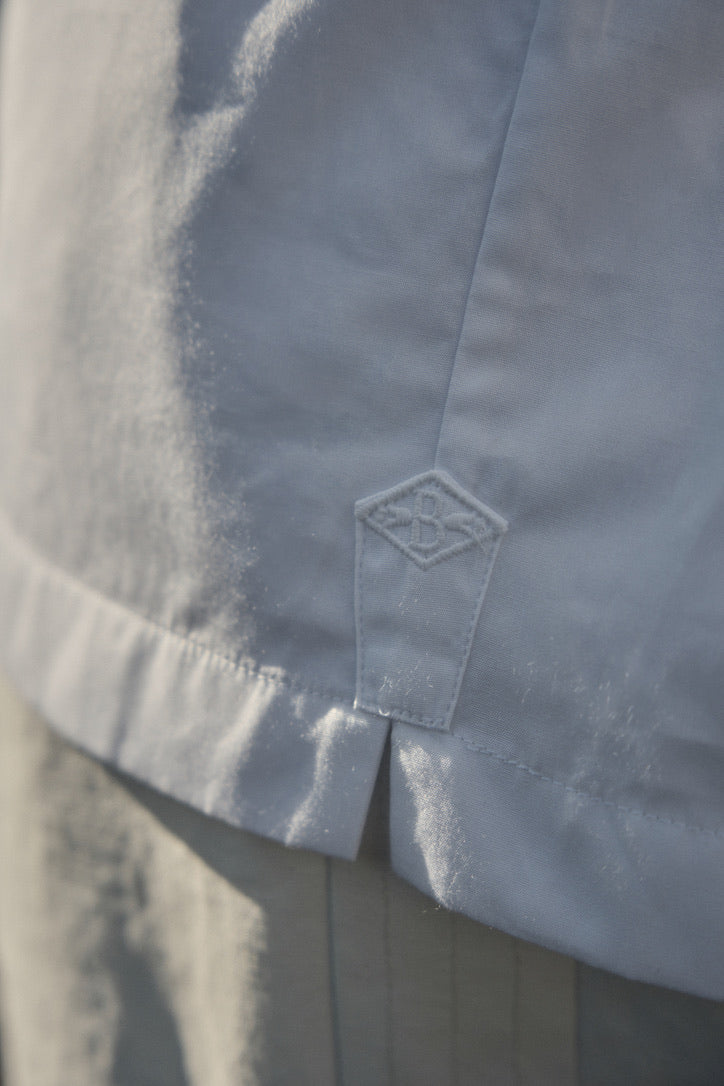 Photo gros plan d'un homme portant une chemise nommé VOYAGE, elle est en 100% Coton, fabriqué en Europe, une coupe Bourrienne, un col Français et des poignets Bourrienne. On voit de près la finition détail du logo bourrienne, brodé en ton sur ton blanc sur la fente de la chemise 