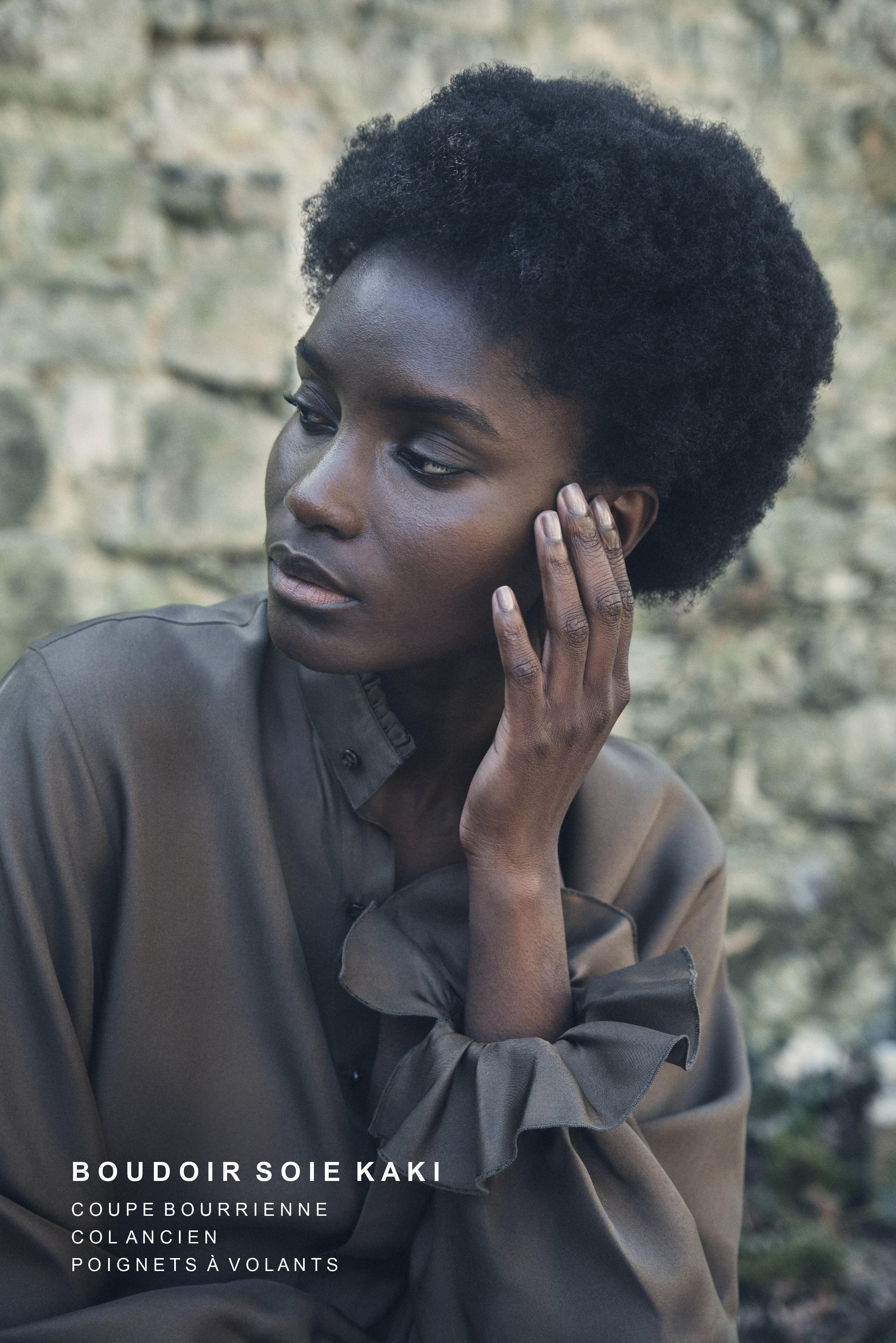 Femme assise portant la chemise BOUDOIR en soie couleur kaki avec poignets à volants, col ancien et coupe fluide, posant devant un mur de pierre.