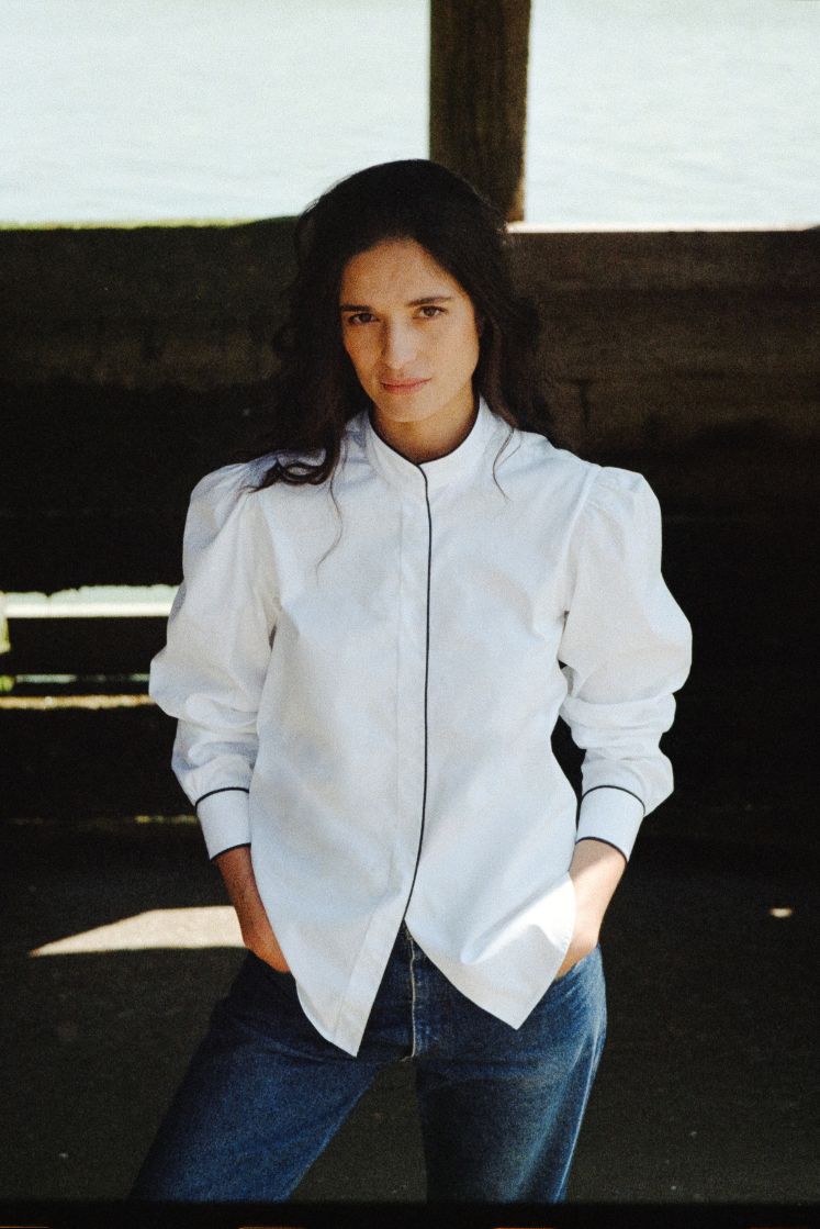 Mannequin vue de face, popeline de coton blanche avec un passepoil bleu marine le long de la gorge de boutonnage ainsi que sur les poignets, mannequin debout avec les mains dans les poches.