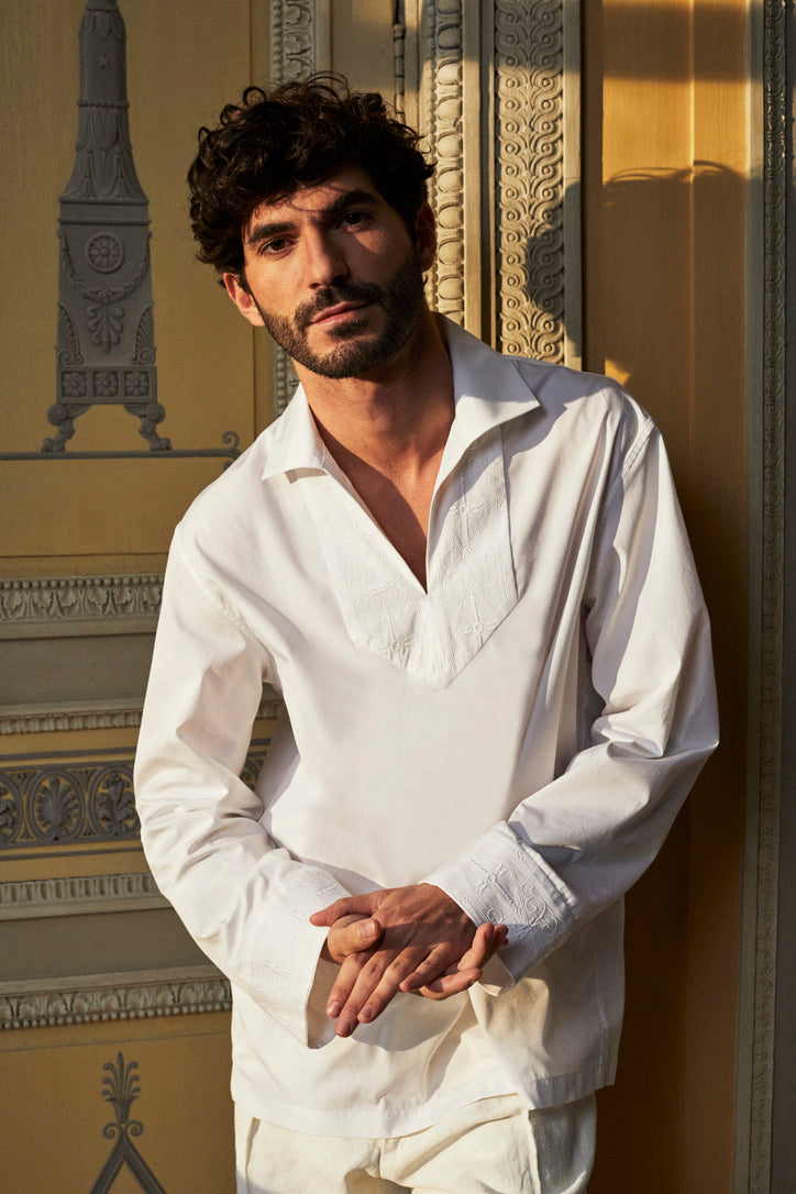 Homme cheveux bruns et bouclés, il porte une chemise blanche avec un col V, la chemise se nomme ARTISAN. La coupe Bourrienne est ample et épurée, des broderies réalisées par un artisan français son présentent sur le col et ses poignets, inspirées par les fresques de l’Hôtel Bourrienne.