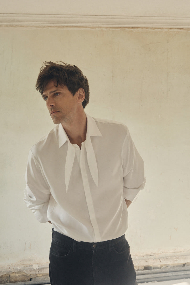 Homme de face buste de trois-quarts, mains croisées dans le dos et regard détourné, portant la chemise PALETTE en soie ivoire à coupe Bourrienne avec un large nœud souple sous le col français. Le tissu satiné, laissé légèrement flottant sur un jean sombre, capte la lumière diffuse qui révèle sa texture lisse et fluide. En arrière-plan, un mur clair et patiné met en valeur le contraste entre la sobriété moderne de la pièce et l’authenticité brute du décor.
