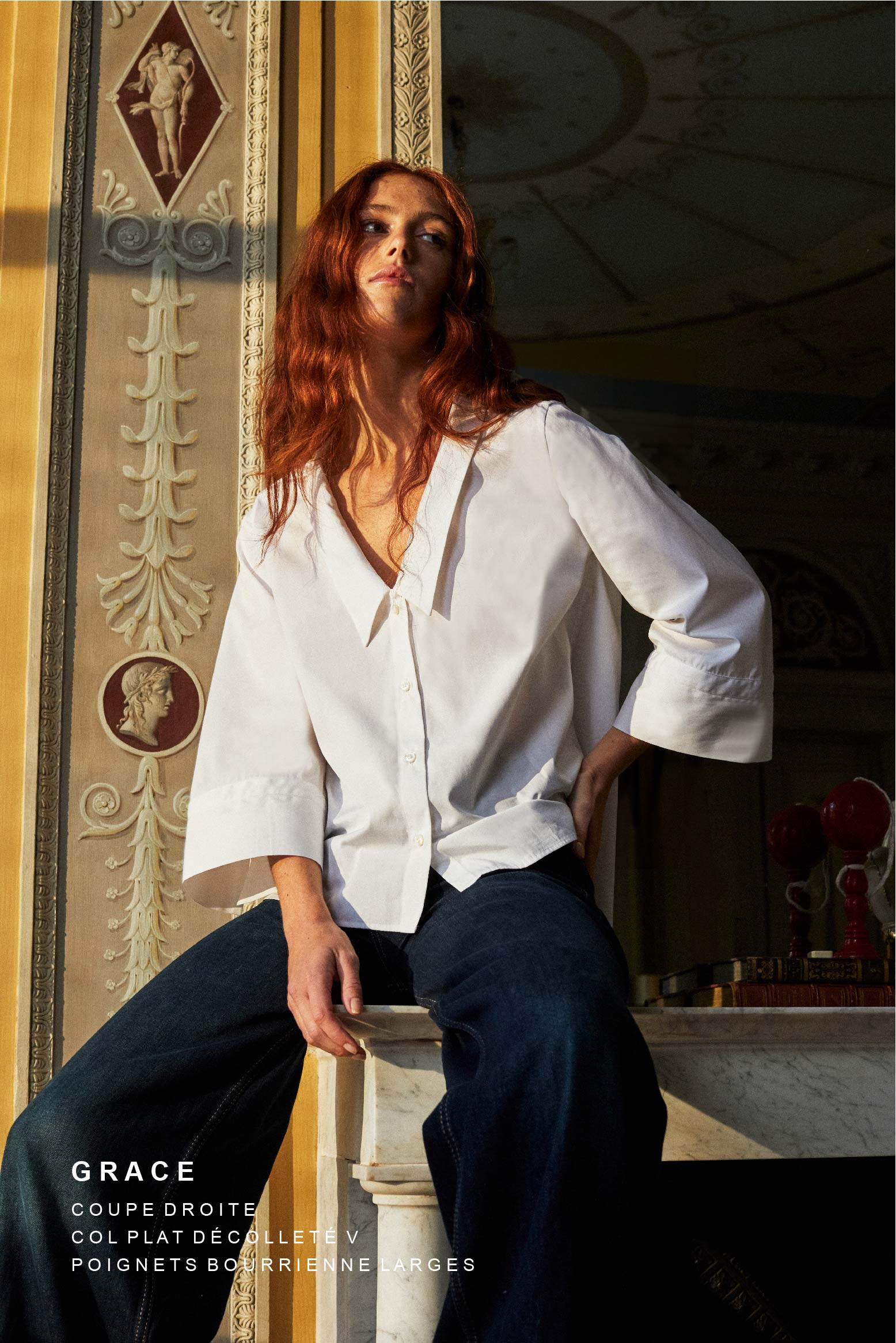 Photo plan américain d'une femme rousse assise sur une cheminée. Elle regarde à sa droite, son bras gauche touche sa hanche. Elle porte une jean droit et une  chemise blanche en popeline double retors italienne Bourrienne Paris X nommé GRACE. Une coupe avec un volume ajusté au niveau du buste et plus évasé en bas, créant une silhouette raffinée. Son col plat et plongeant ajoute une touche de féminité. Des manches ¾ avec des details brodés. 