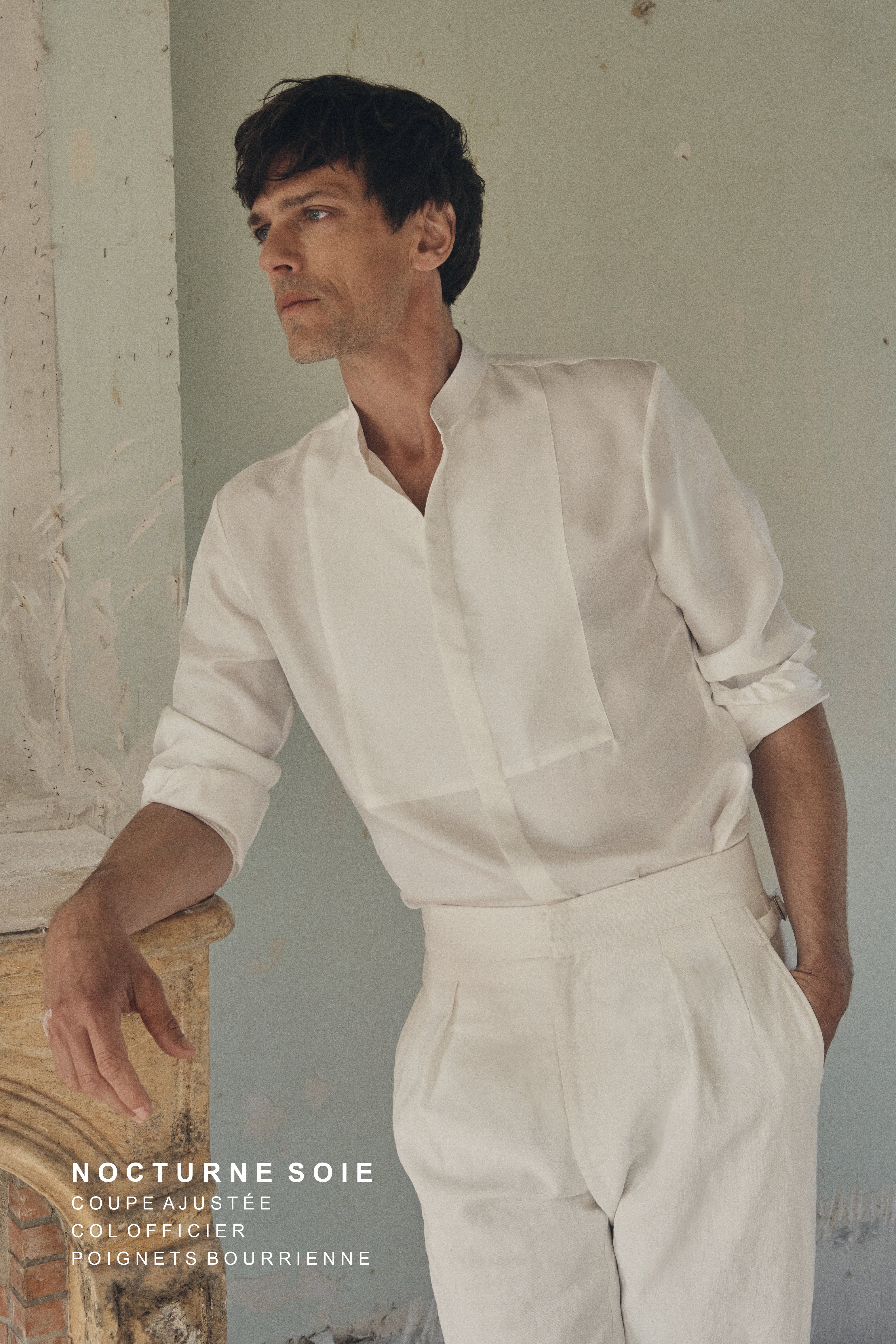 Homme de profil trois-quarts, appuyé contre un manteau de cheminée en pierre patinée, portant une chemise NOCTURNE en soie blanche à coupe ajustée, col officier et plastron revisité. Les manches sont retroussées au-dessus du poignet, dévoilant les poignets Bourrienne signature, et le plastron en nid d’abeille ajoute un relief subtil sur la poitrine. Le décor minimaliste aux murs clairsemés et la lumière naturelle mettent en valeur la fluidité et la finesse du tissu.