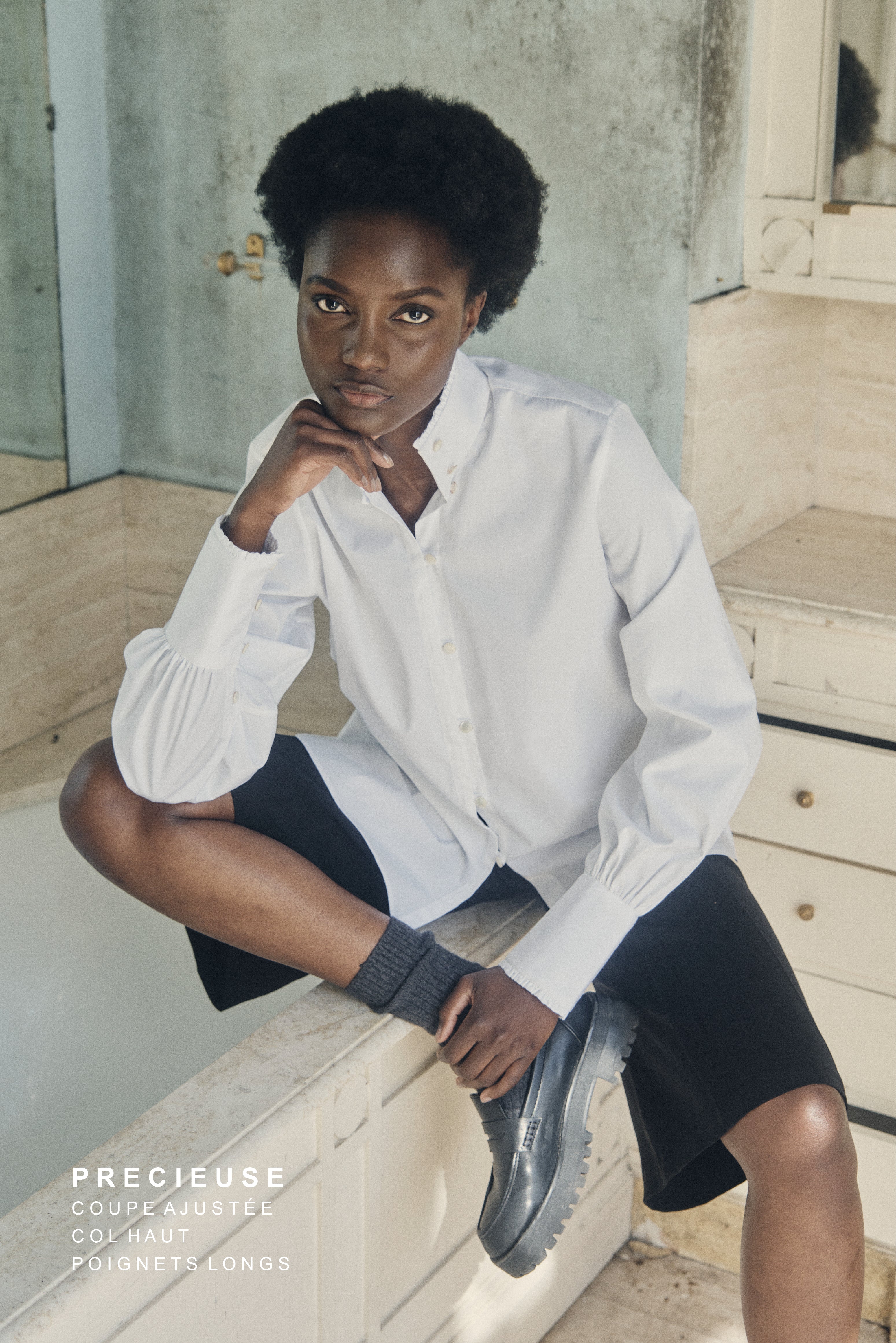 Femme assise sur une baignoire portant la chemise blanche PRÉCIEUSE, un bermuda noir, une paire de mocassins noire et des chaussettes grises légèrement montantes.