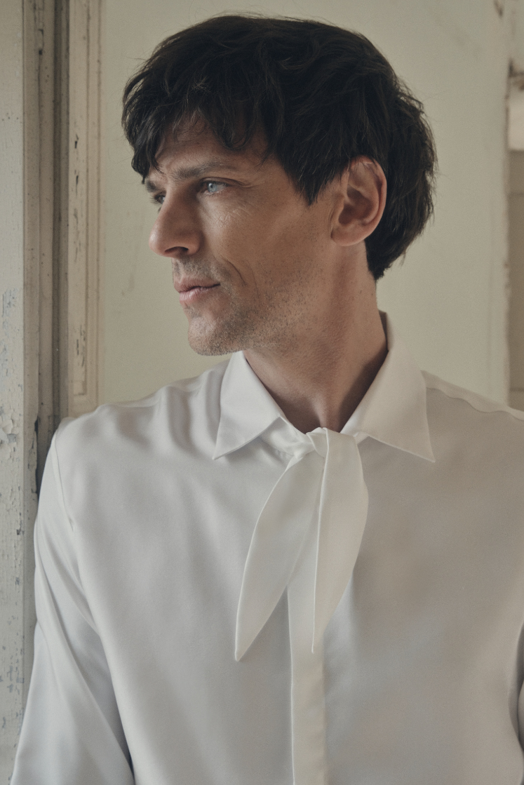 Homme de profil, appuyé contre l’embrasure d’une fenêtre, portant la chemise PALETTE en soie blanche à coupe Bourrienne et col français, dont le large nœud décontracté apporte une touche de fluidité. Le décolleté ouvert et le tombé souple du tissu mettent en valeur l’éclat satiné de la soie, tandis que les poignets classiques ajoutent une note raffinée. La lumière douce qui filtre par la fenêtre souligne la texture délicate du vêtement et le contraste entre l’élégance moderne et le décor patiné.