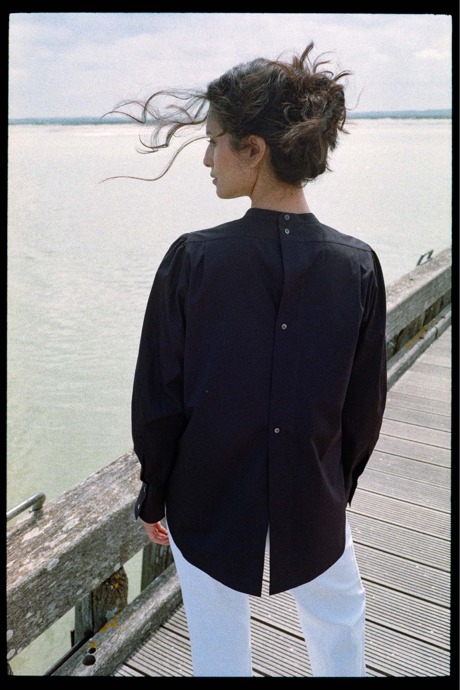 BOURRIENNE PARIS X: Photo prise de dos, le mannequin regarde à gauche et elle a des mèches volantes. La photo nous permet de voir les détails des 4 boutons dans le dos.