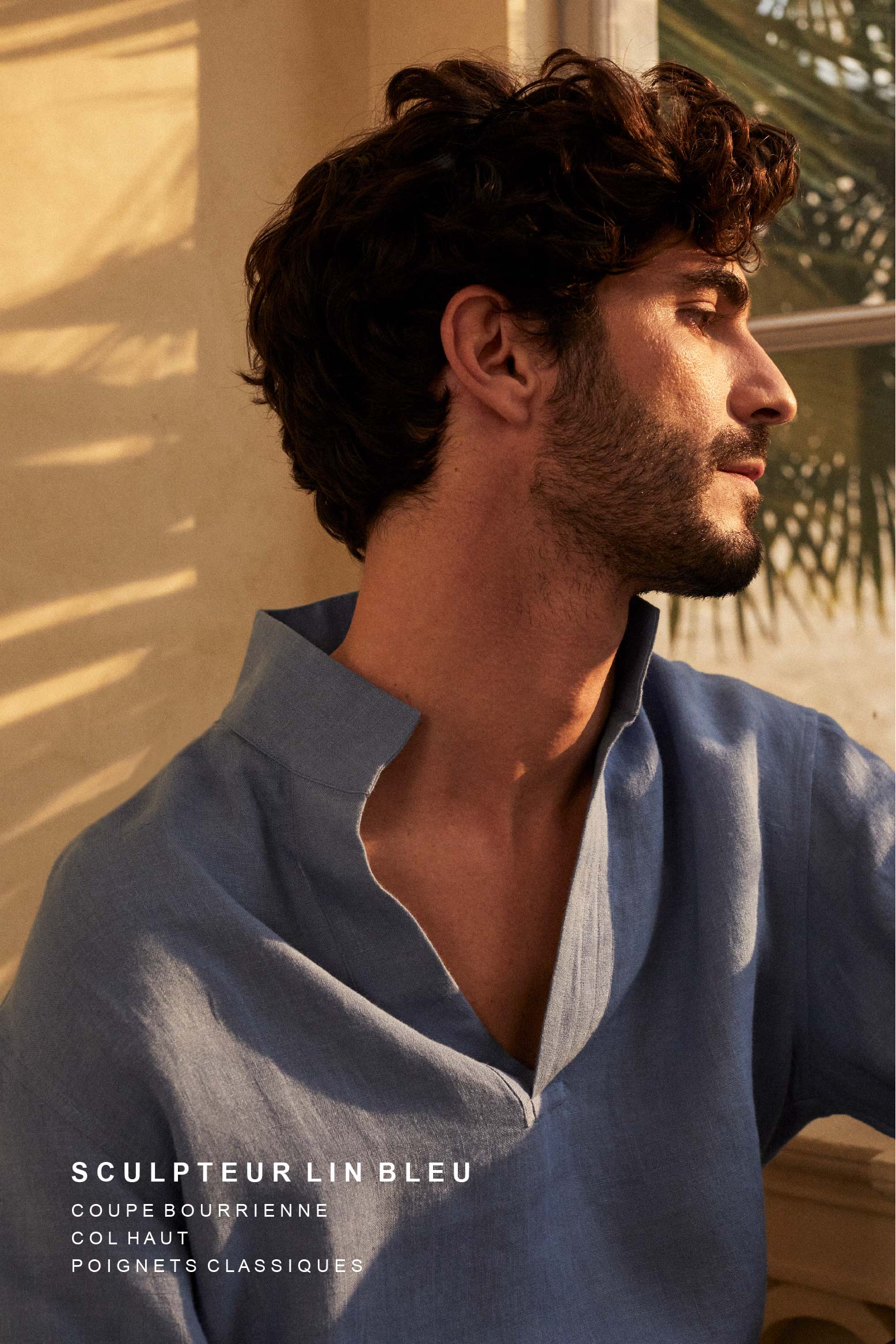 Homme brun portant la chemise SCULPTEUR en lin bleu Bourrienne Paris X, close-up sur le col haut élégant. Assis près d'une fenêtre avec des palmiers en arrière-plan, baigné par une lumière dorée et naturelle.