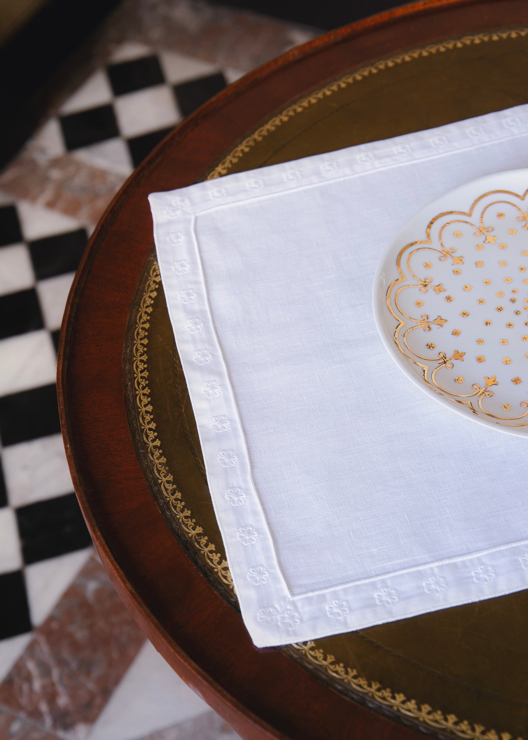 Set de table blanc orné d'un galon brodé reprenant la fleur de l'Hôtel de Bourrienne, posé sur une table en bois. L'assiette issue de la même collection est posée sur le set de table.