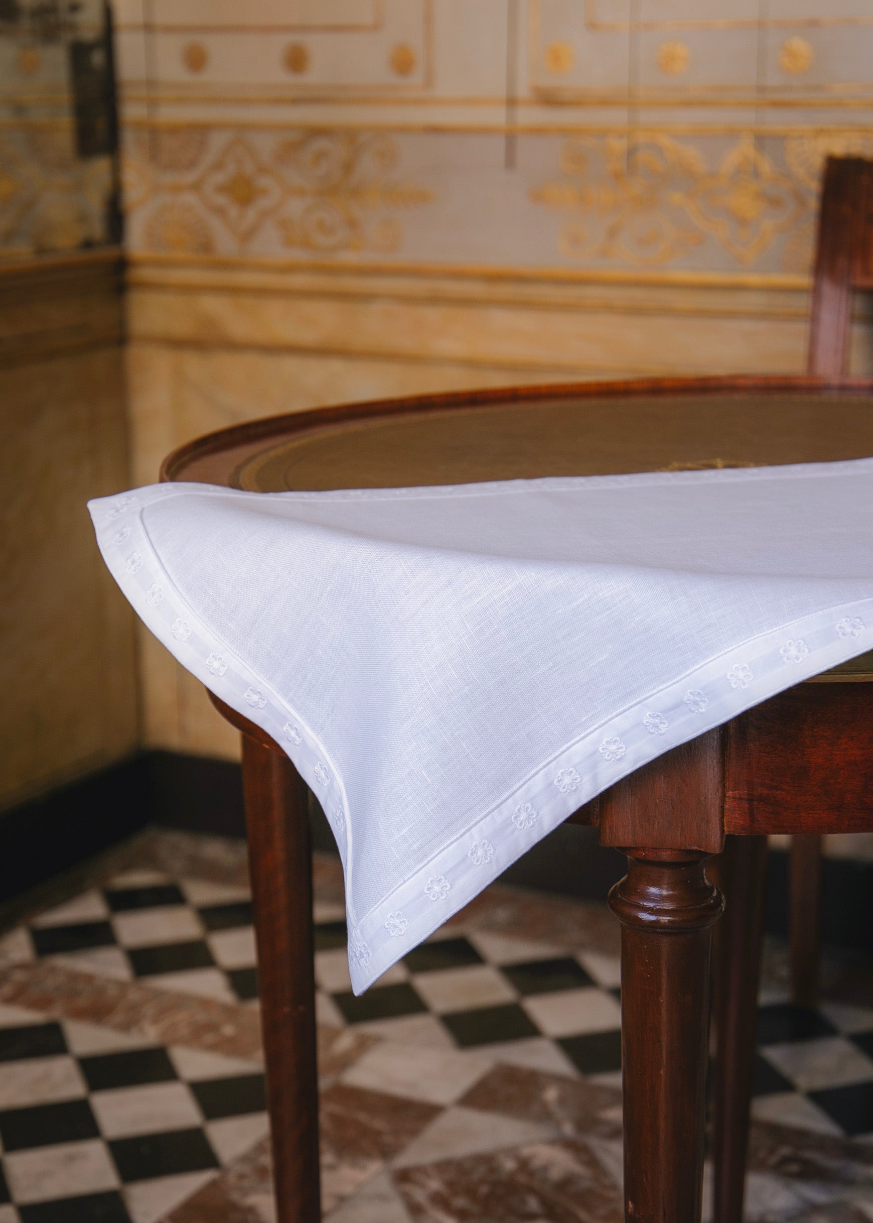 Set de table blanc orné d'un galon brodé ton sur ton, reprenant la fleur de l'Hôtel de Bourrienne, posé sur une table en bois ronde dans l'Hôtel de Bourrienne.