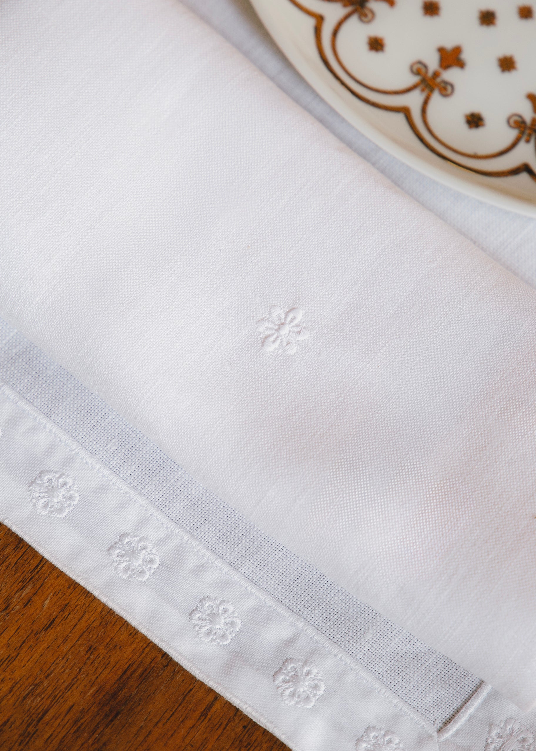 Close-up sur le set de table blanc et son galon brodé ton sur ton. La serviette blanche issue de la même collection est posée sur le set et reprend également la broderie de la fleur de l'Hôtel de Bourrienne.