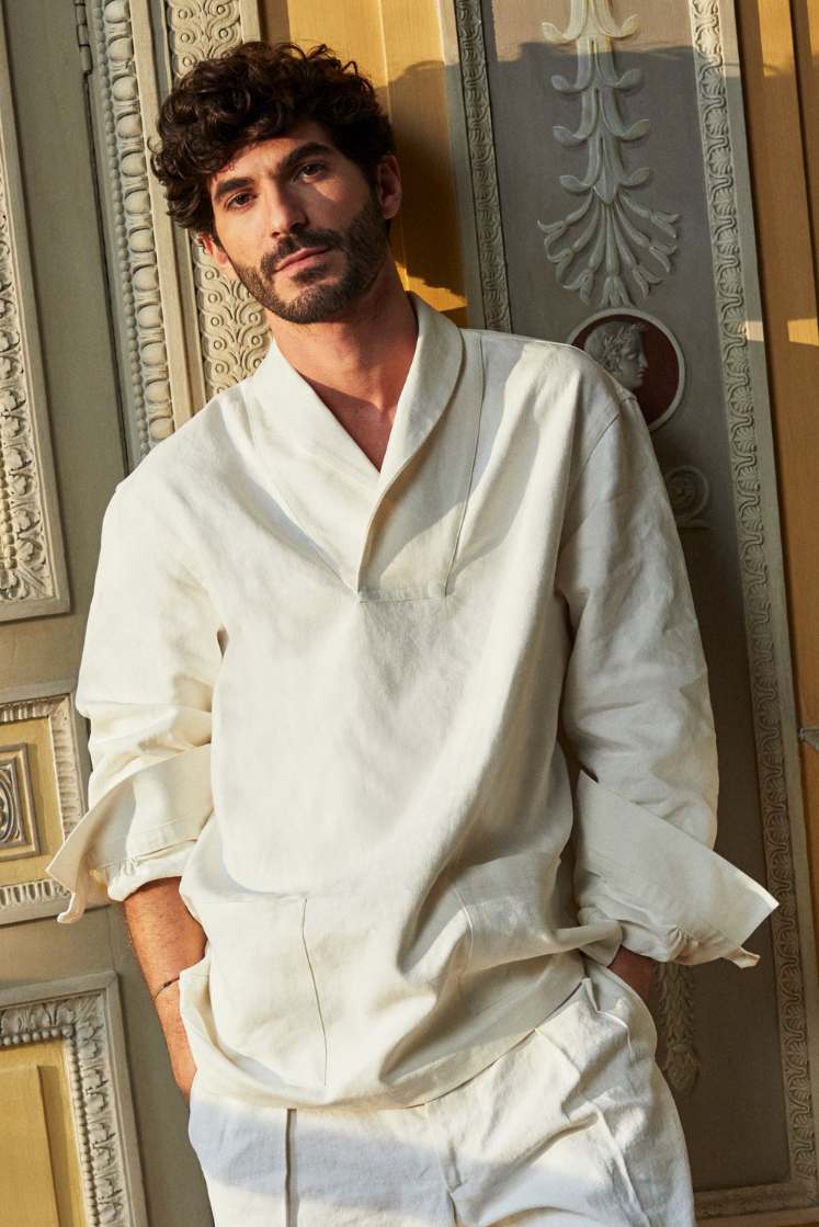 Homme brun adossé contre un mur, portant une chemise col châle en lin blanc signée Bourrienne Paris X, posant dans l'Hotêl de Bourrienne, les mais dans les poches de son pantalon, de face.