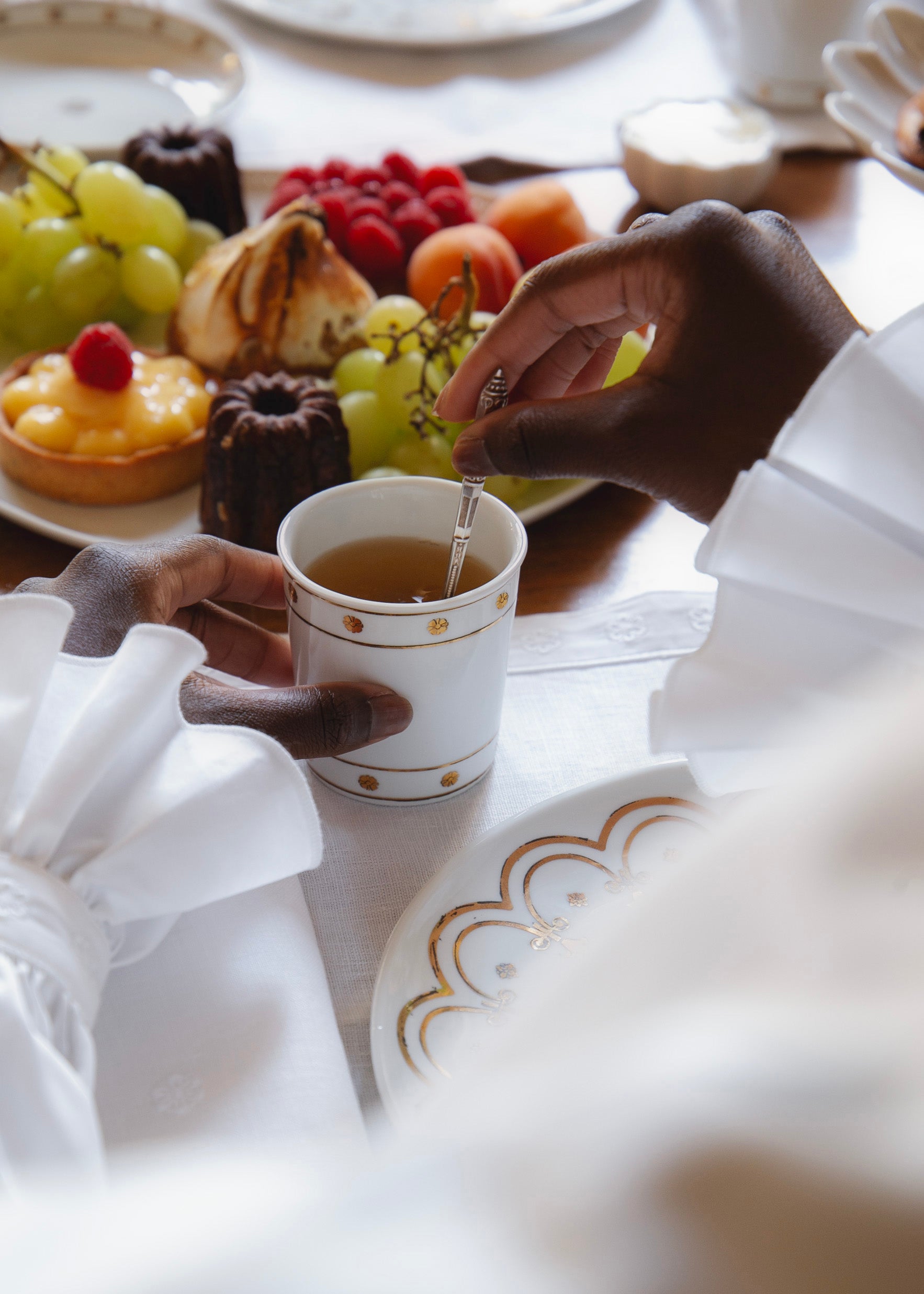 Une personne vêtue d’une chemise blanche à poignets volantés mélange son thé avec une petite cuillère argentées dans la timbale en porcelaine blanche ornée de motifs dorés. À côté, un plateau de pâtisseries et raisins complètent la scène sur une table dressée.