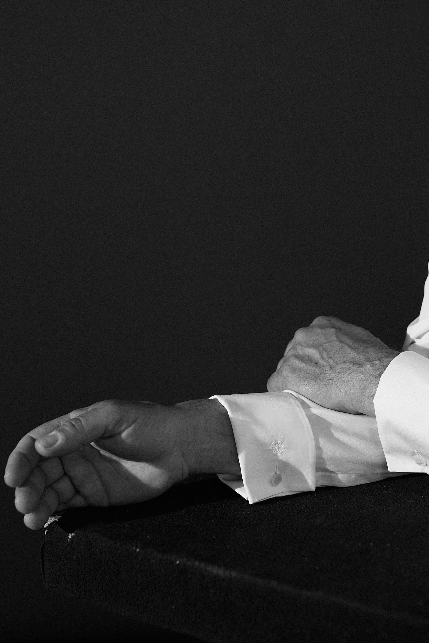 Photo gros plan noir et blanc. On voit une main posée sur une table en bois qui croise le deuxième bras également  posé sur la table. C'est les bras d'un homme portant une chemise blanche avec des poignets mousquetaires. 