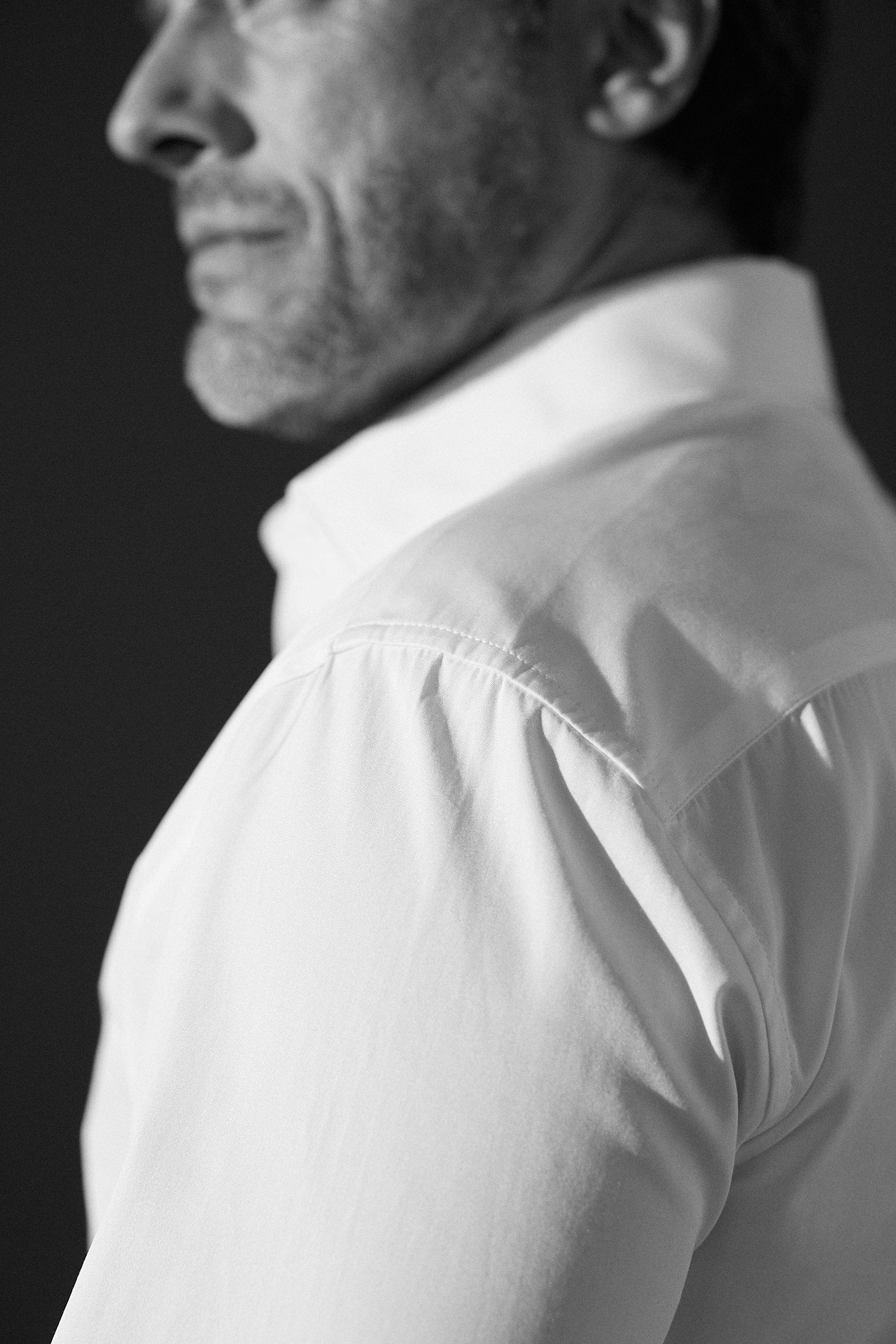 Photo en noir et blanc, plan poitrine. Un homme porte une chemise blanche en popeline double retors, il se tient de profil on voit au deuxième plan son épaule gauche et le détail de l'épaule de la chemise qui fronce légèrement et au troisième plan le col italien.  