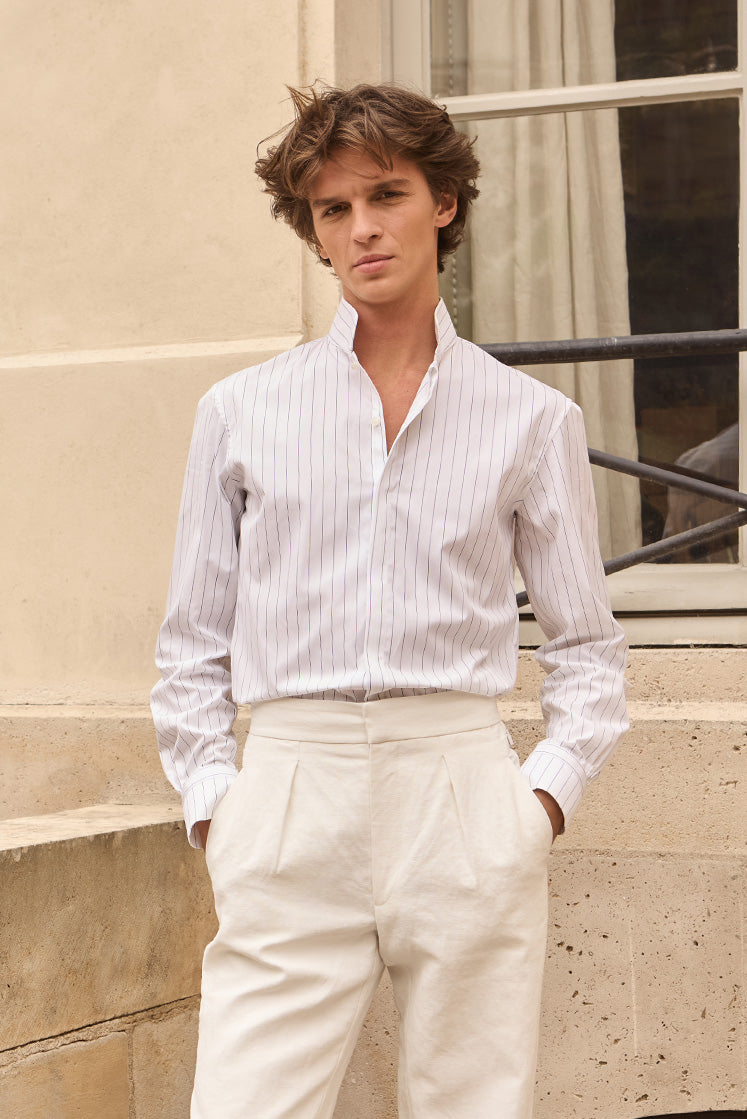 Homme dans une cour portant une chemise blanche rayée bleu marine à col haut, rentrée dans un pantalon en lin.