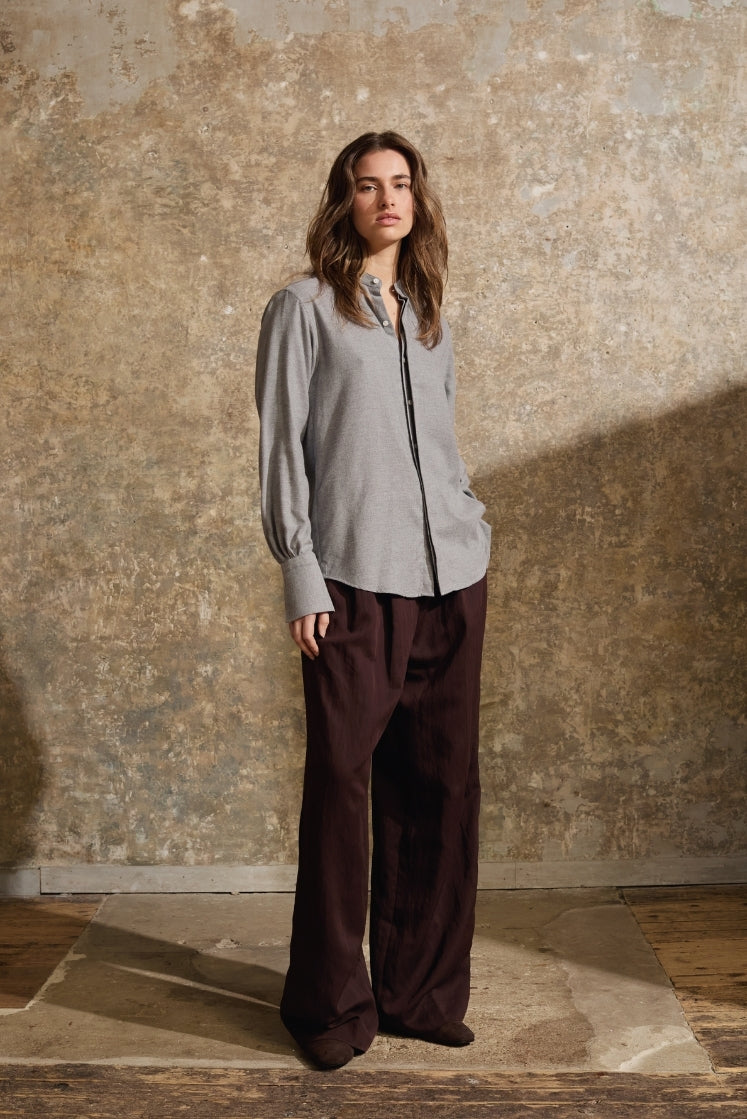 Femme portant chemise Astrale en flanelle grise boutonnée, pantalon marron large, pose élégante devant mur texturé